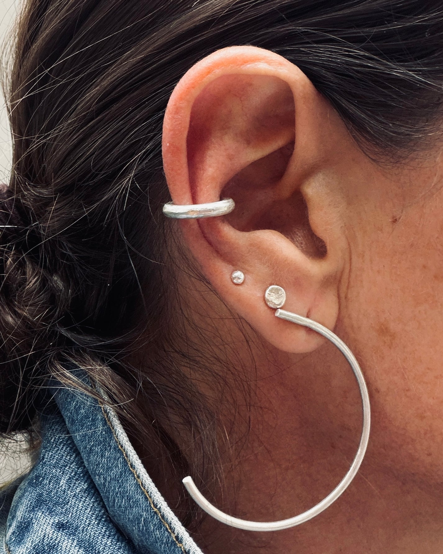 A close-up of a person's ear showcasing a silver ear cuff, a small stud earring, and a larger hoop earring.