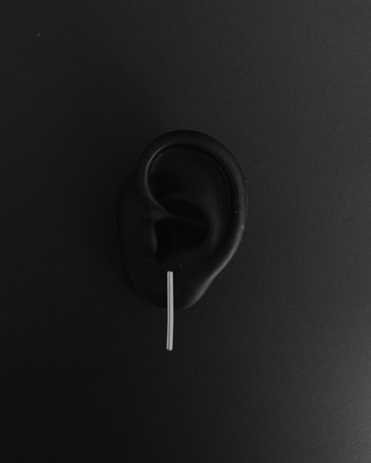 A black silicone ear plug with a thin silver metal bar inserted into the ear canal, shown against a dark gray background.