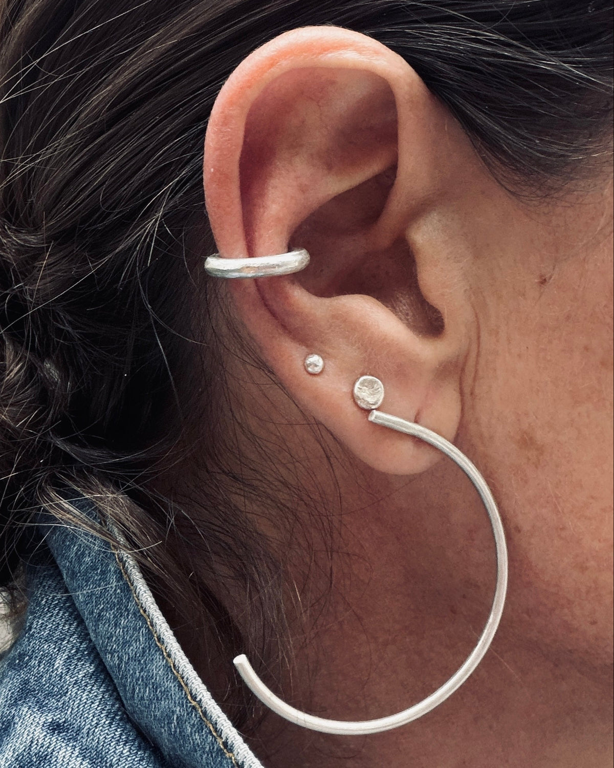 A close-up of a person's ear showcasing a silver ear cuff, a small stud earring, and a large silver hoop earring.