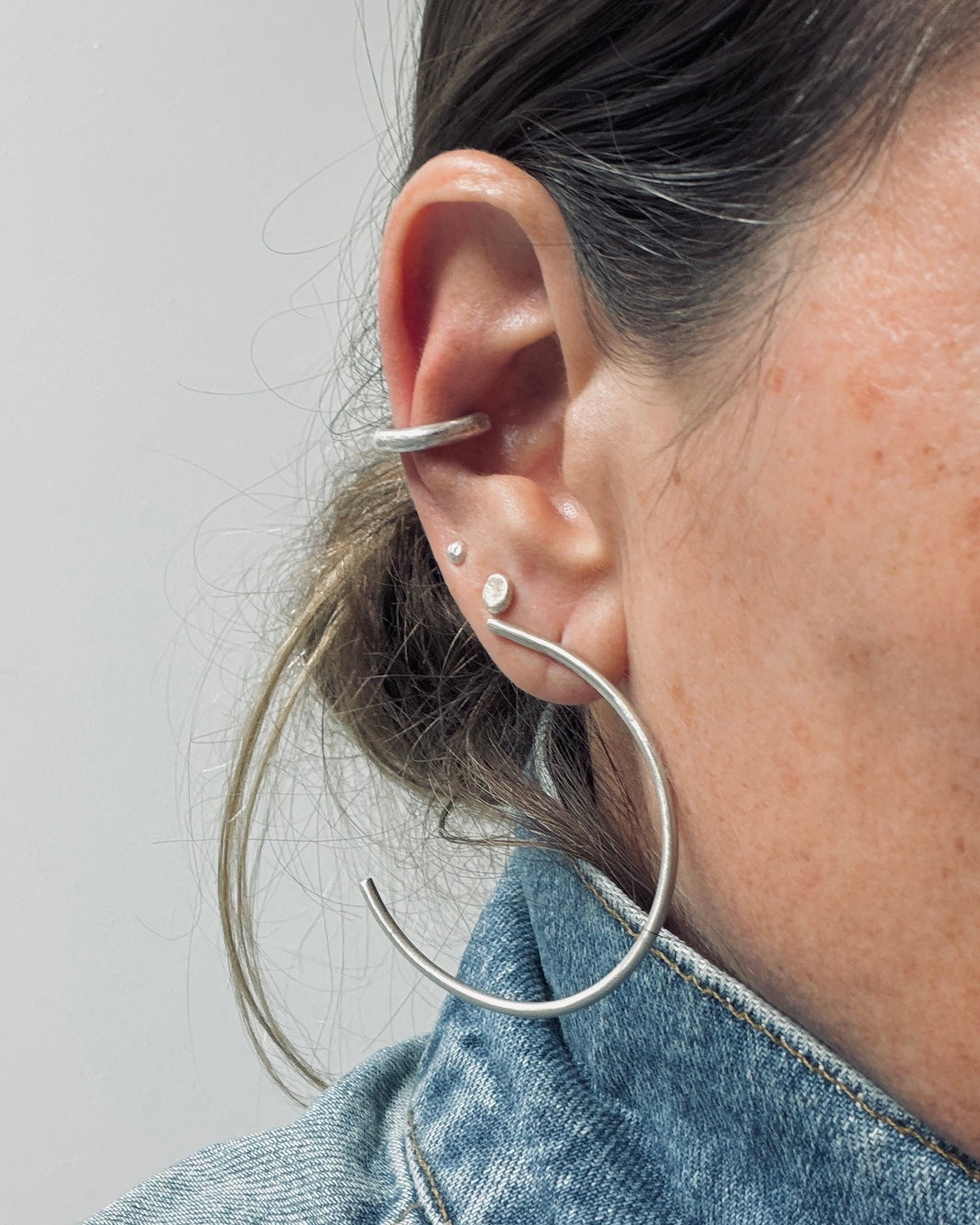 Close-up of a person's ear wearing multiple silver earrings, including a large hoop earring, a curved ear cuff, and two small stud earrings, against a plain light gray background.