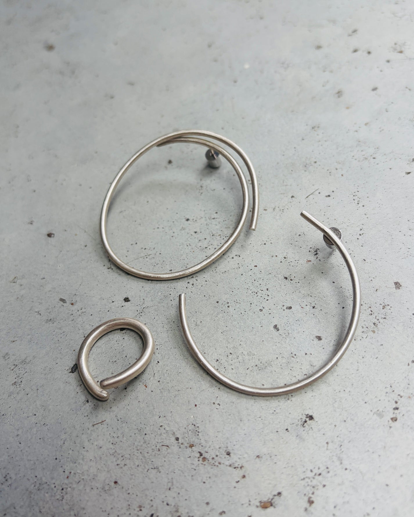 Three minimalist silver wire hoop earrings in different sizes arranged on a textured gray concrete surface.