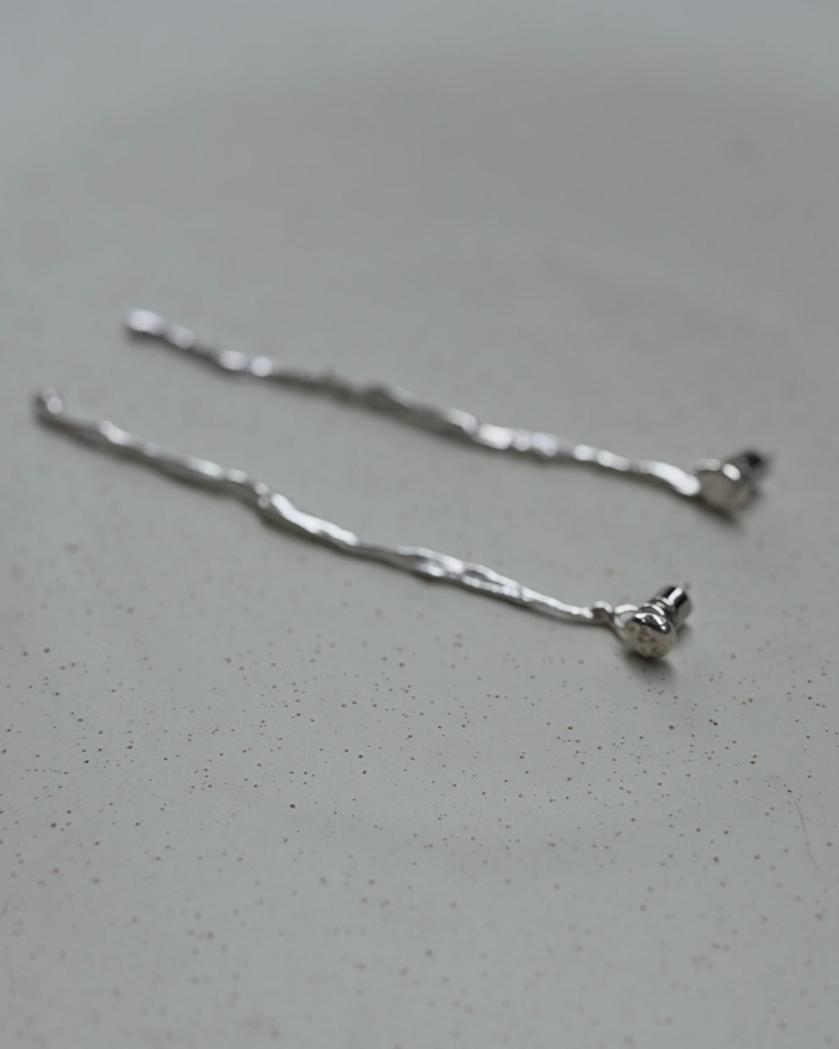 A pair of delicate silver bobby pins with a textured, hammered finish, resting on a light gray speckled ceramic surface.