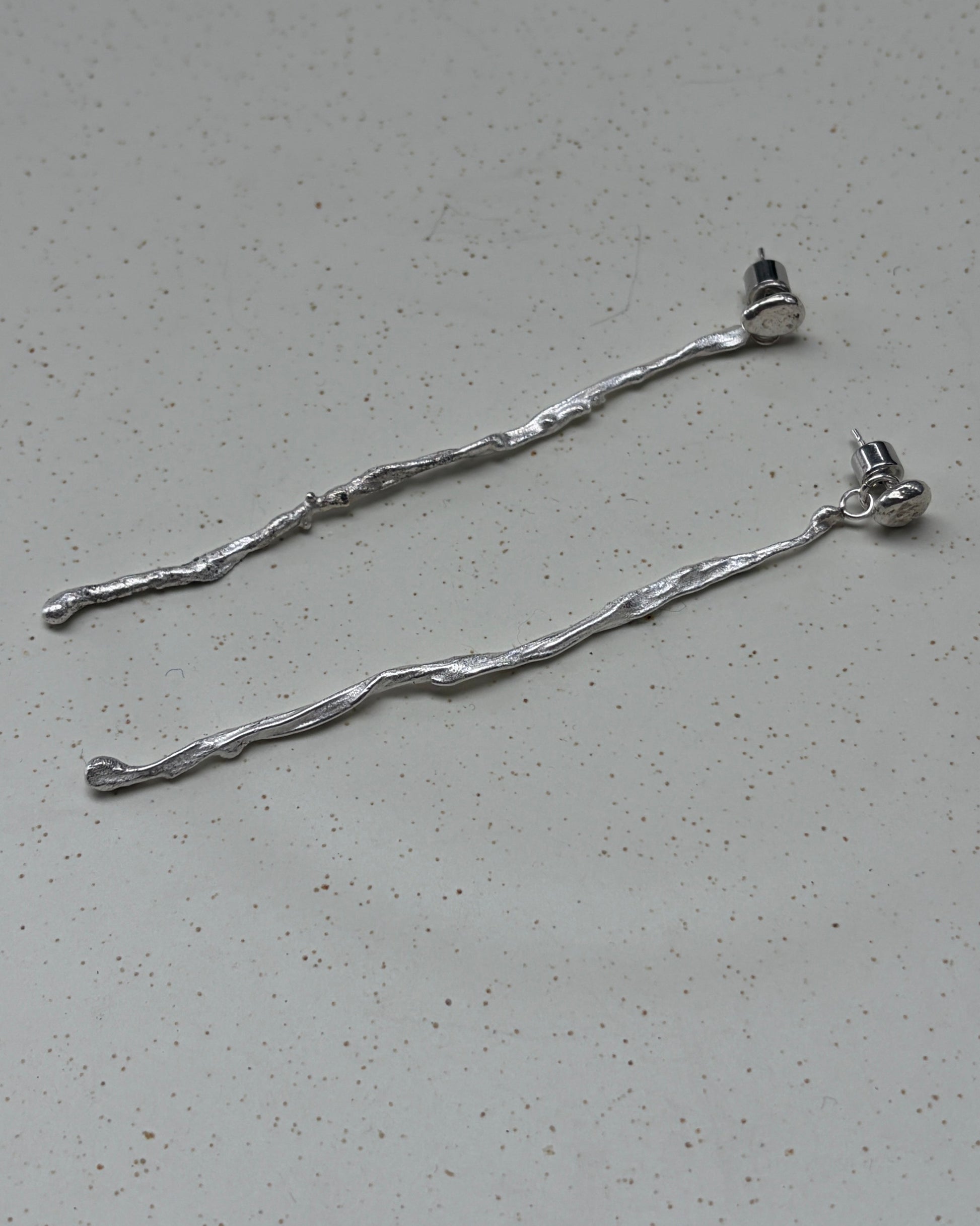 A pair of long, twisted silver metal hair pins or extension bars lying on a speckled light gray surface.