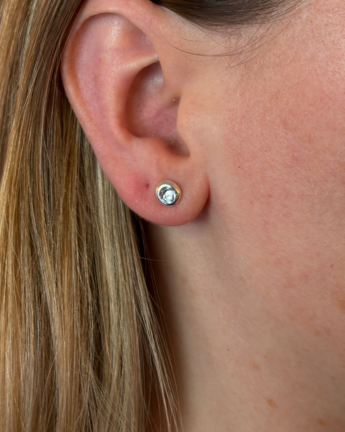 A close-up of a person's ear wearing a small silver stud earring with a clear gemstone, shown against skin and blonde hair.