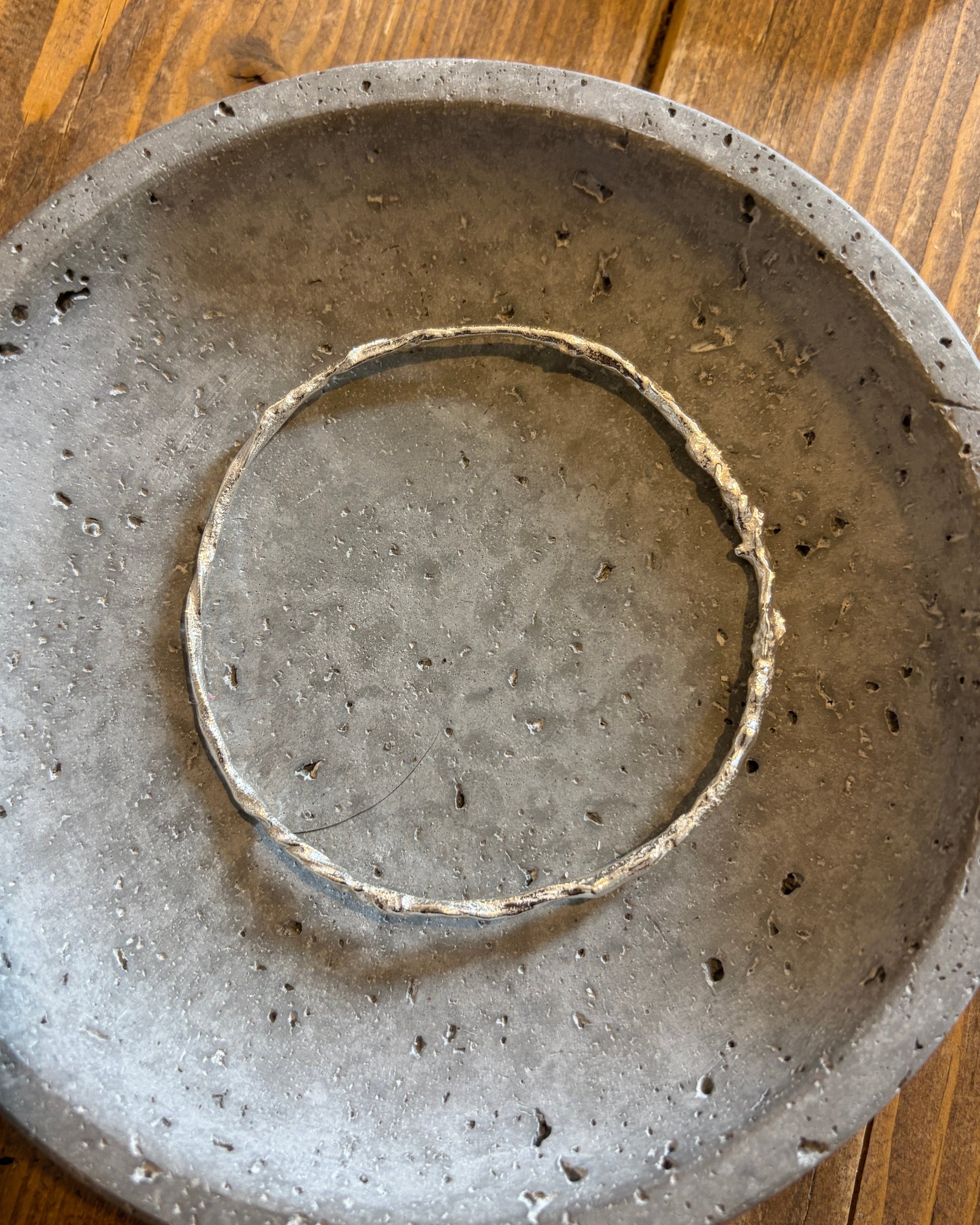 A handmade circular concrete tray with a textured, speckled gray finish, featuring a smaller concentric ring cut into the center, displayed on a wooden surface.