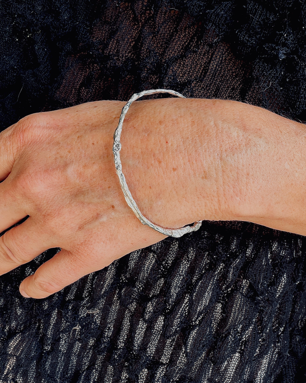 A close-up of a person's wrist wearing a thin, textured silver bangle bracelet against a black lace fabric background.