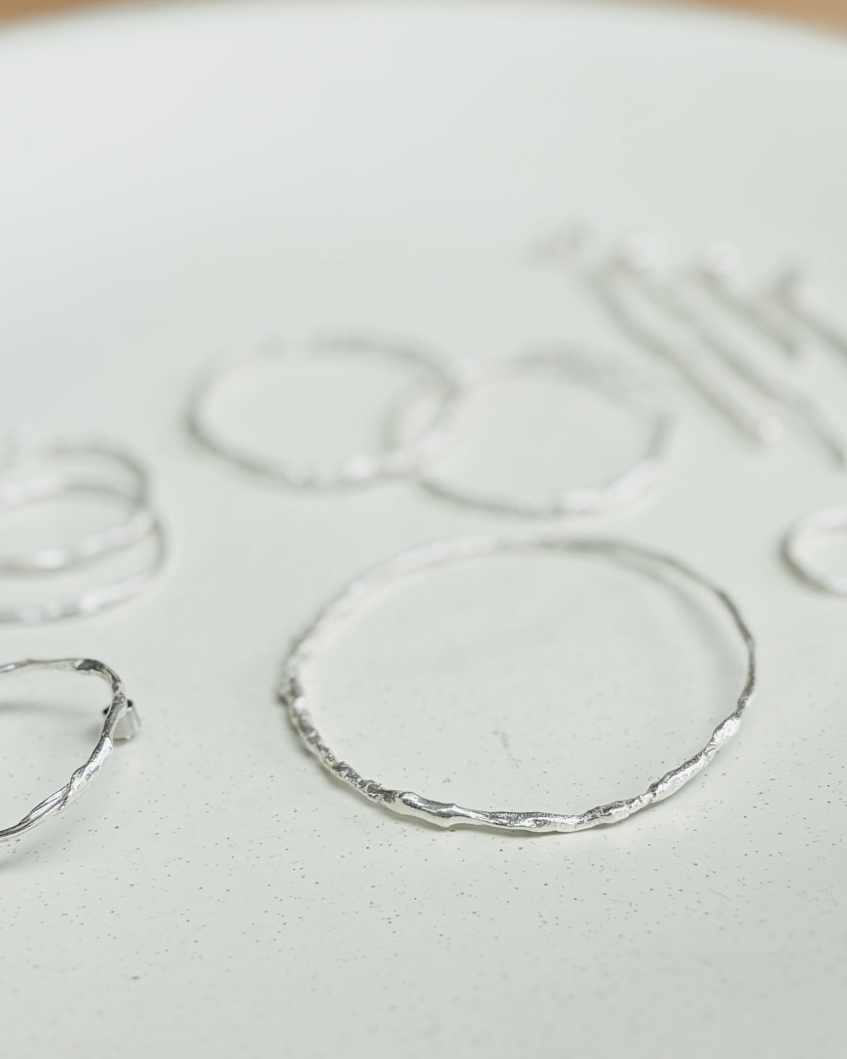 A close-up of several thin silver rings with a hammered or textured finish, arranged on a light off-white surface.
