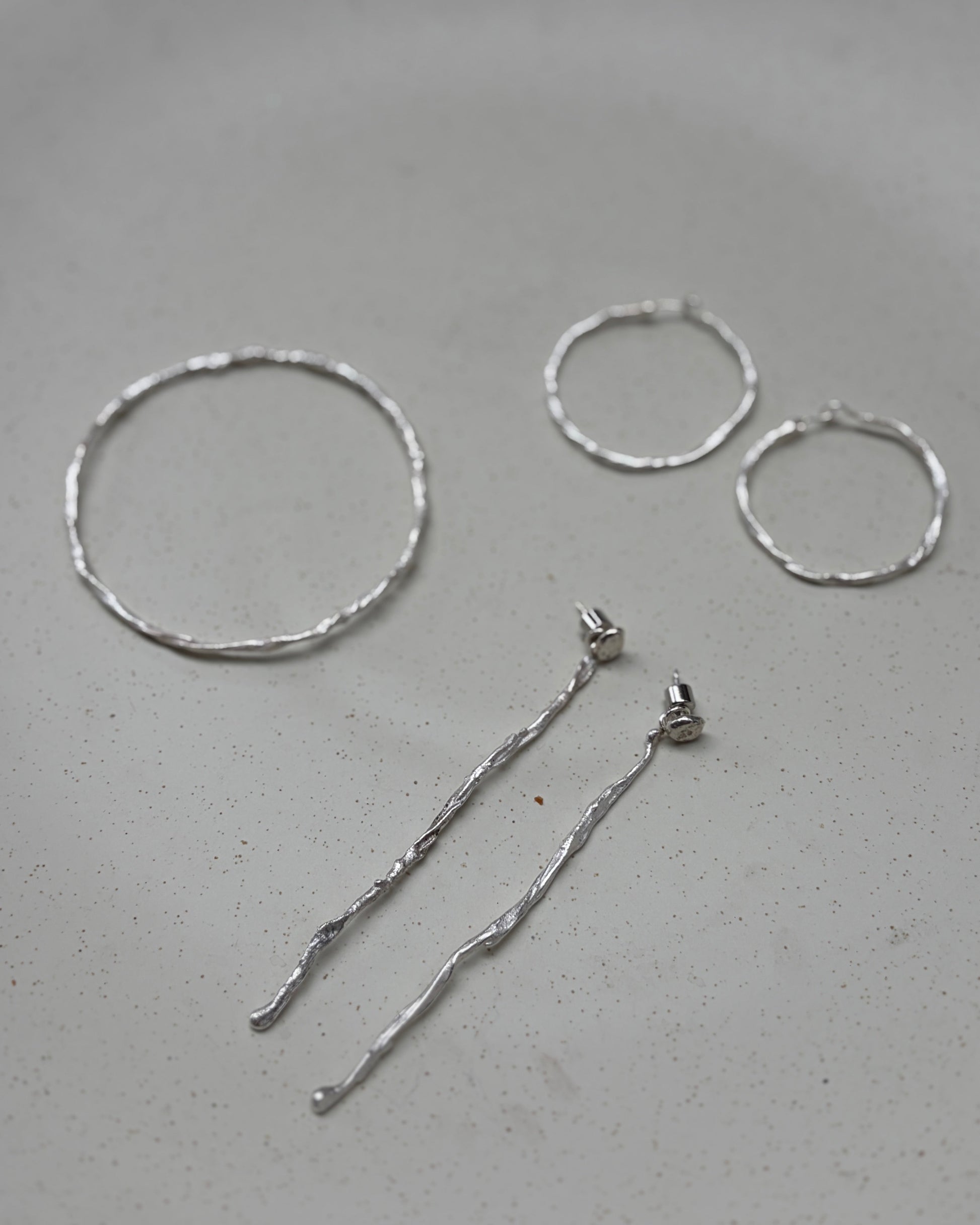 A set of silver-toned jewelry pieces including a textured circular hoop, two smaller textured hoop earrings, and two long textured dangle earrings, arranged on a speckled light gray ceramic plate.