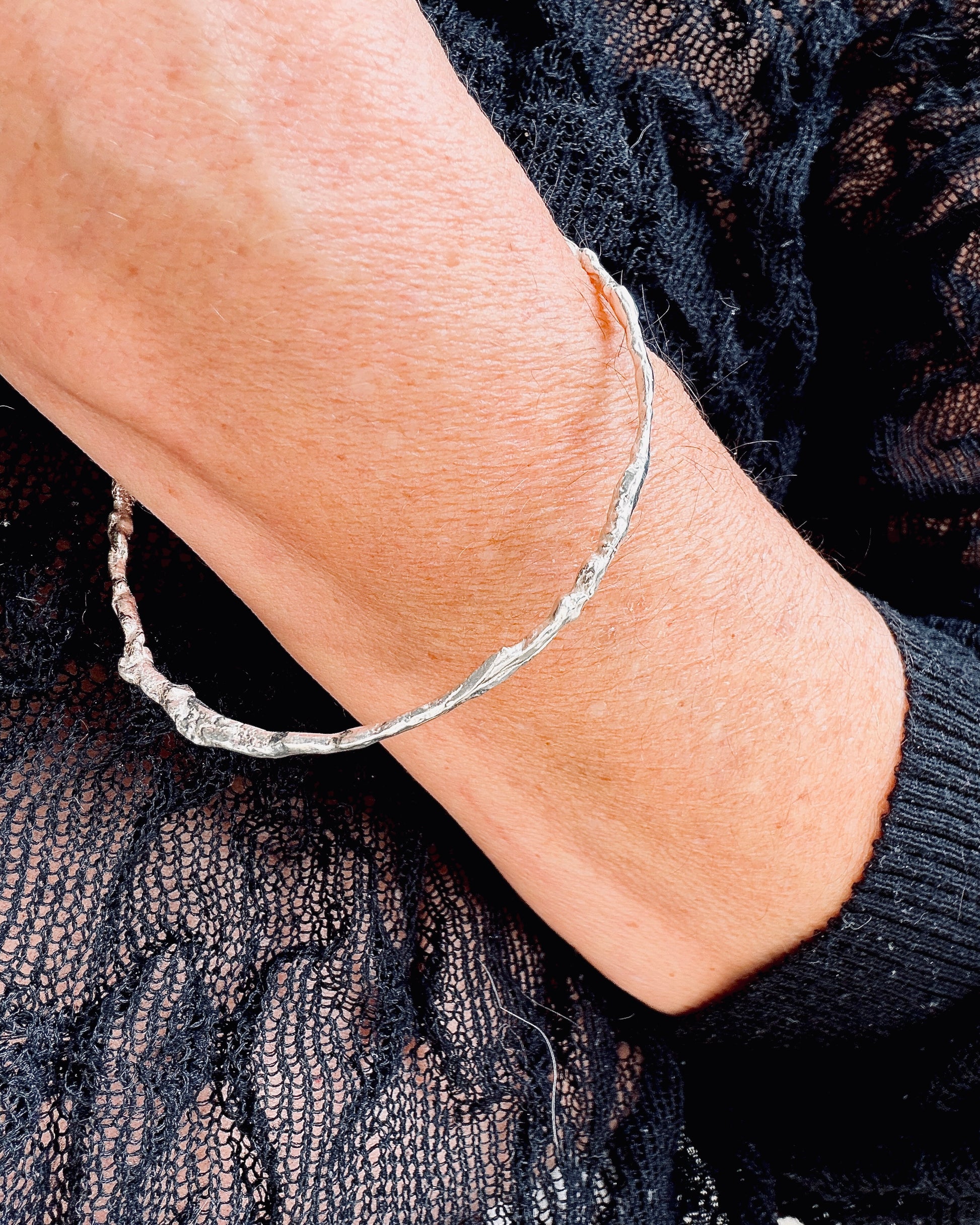 A close-up of a person's wrist wearing a thin, textured silver bangle bracelet, paired with a black lace top.