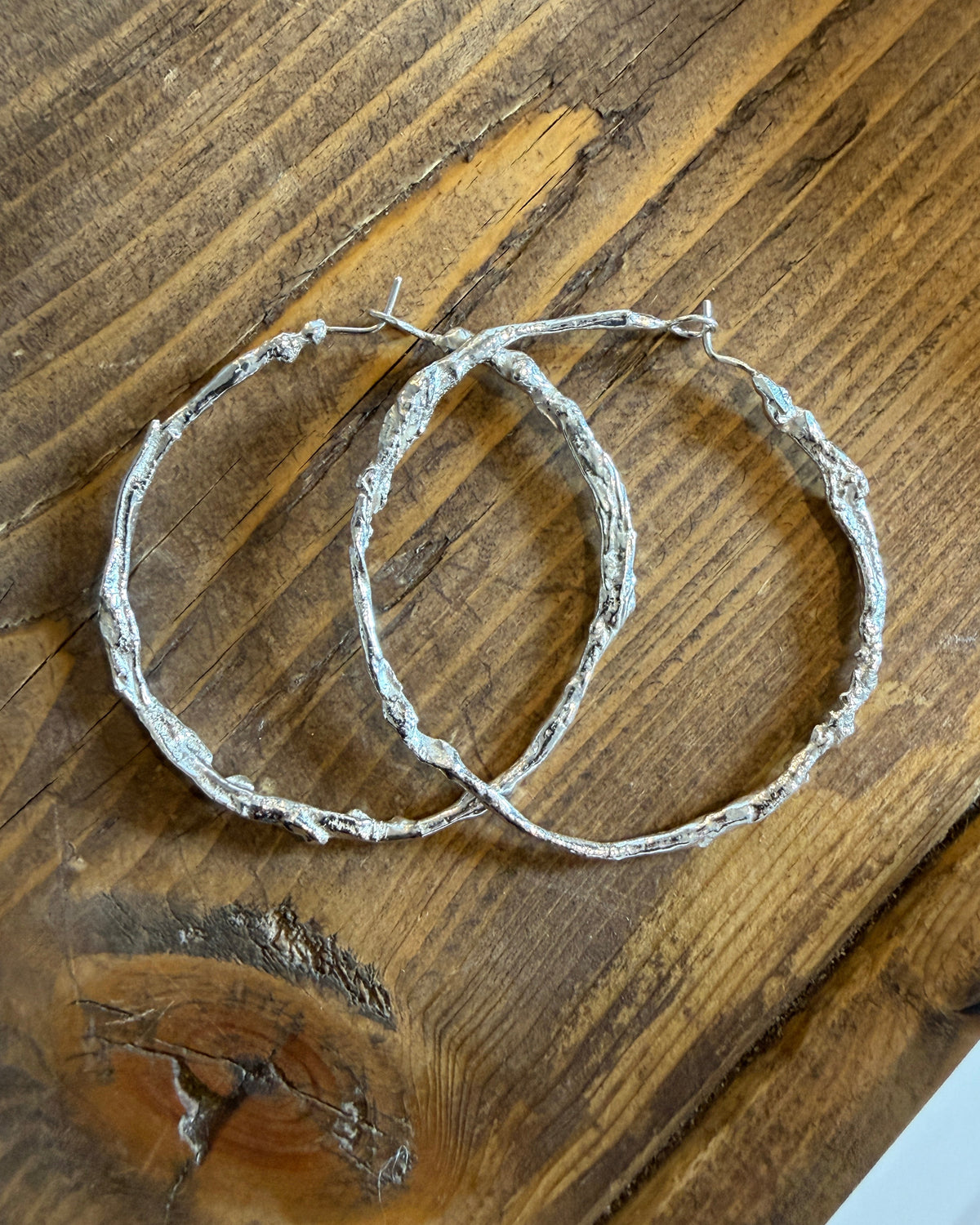 Two rustic silver wire hoop earrings with a rough, textured finish, displayed on a dark brown wooden surface.