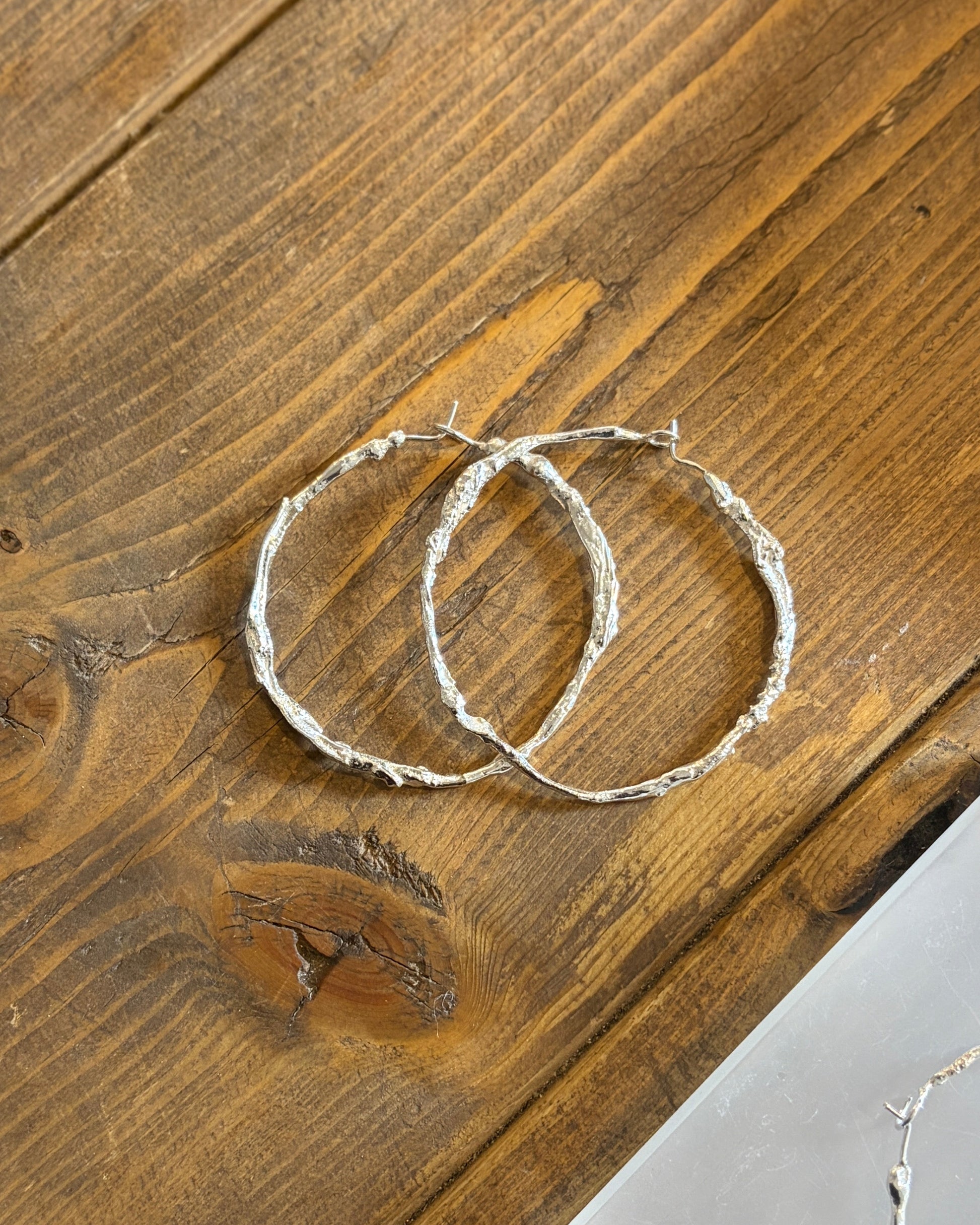 Three textured silver hoop earrings displayed on a rustic wooden table, with one pair resting on a clear acrylic stand.
