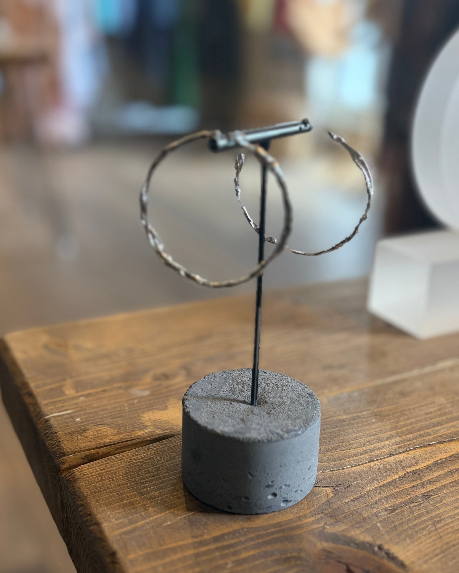 A pair of hammered silver hoop earrings displayed on a black metal stand with a round gray concrete base, set on a rustic wooden table.