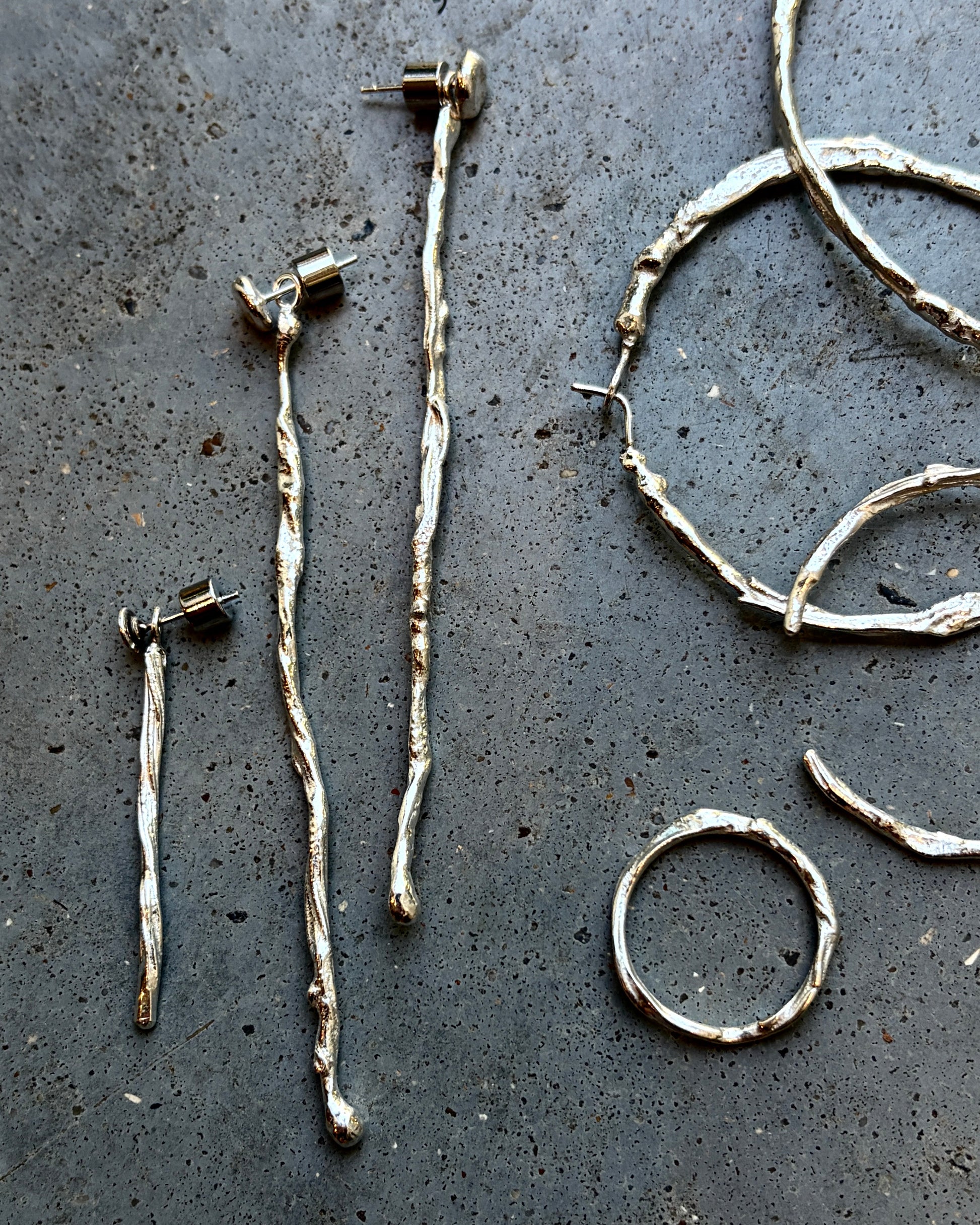 Handmade twig-style silver jewelry pieces laid out on a dark textured surface, including several long earrings, a ring, and a curved cuff.
