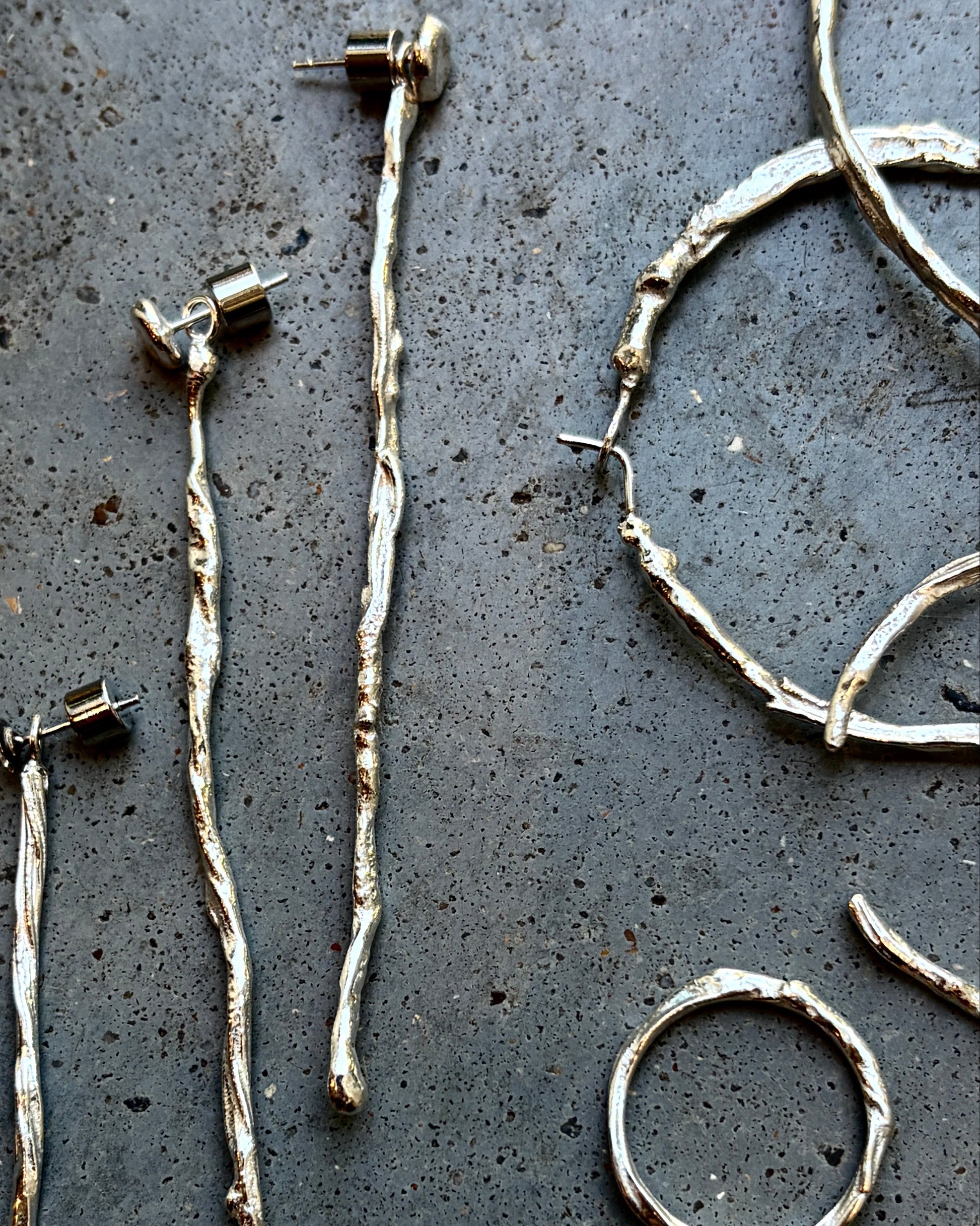 Handmade twig-style silver jewelry pieces laid out on a dark textured surface, including several long earrings, a ring, and a curved cuff.