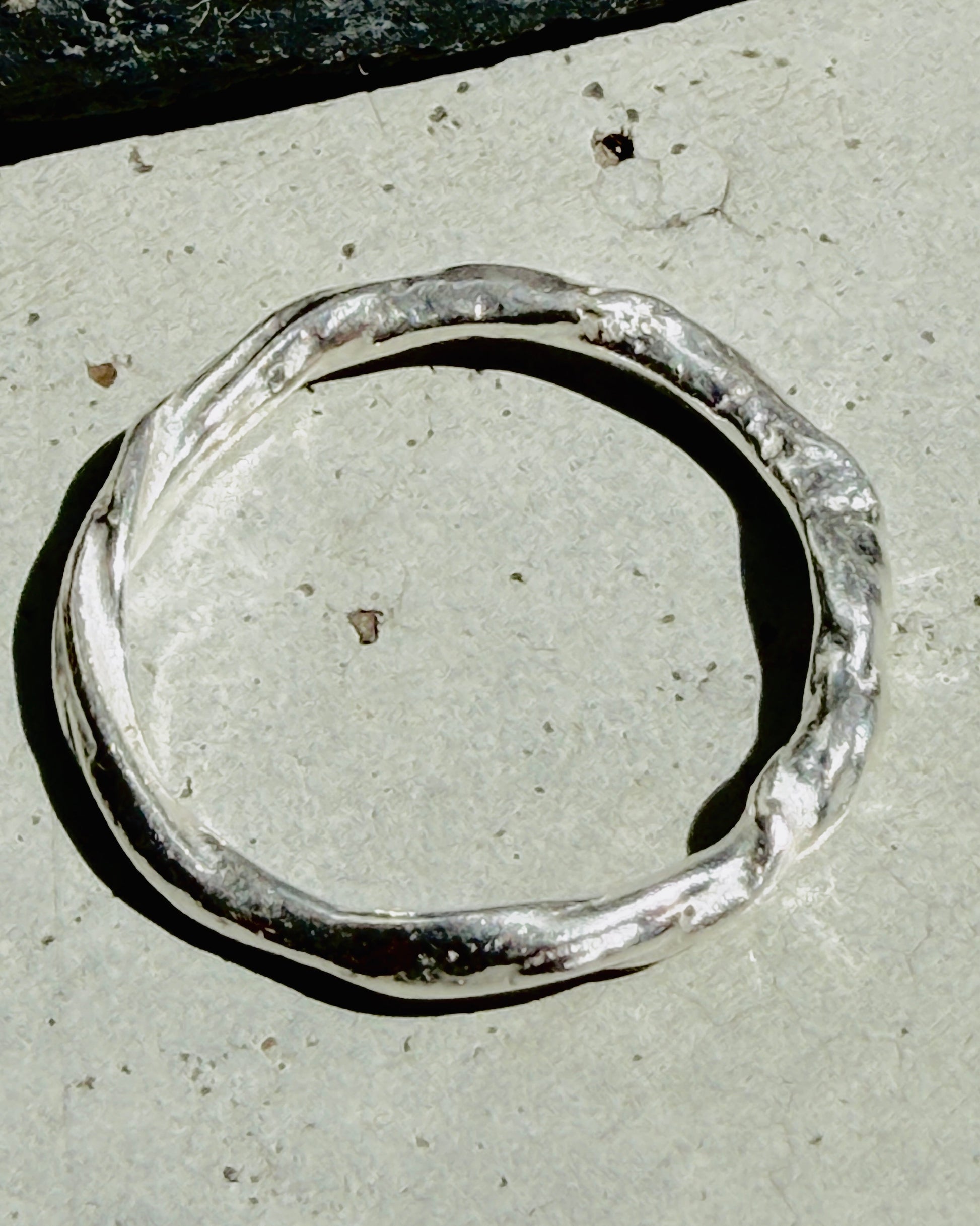 A hammered silver-toned metal ring with an organic, wavy shape, photographed on a light gray concrete surface.