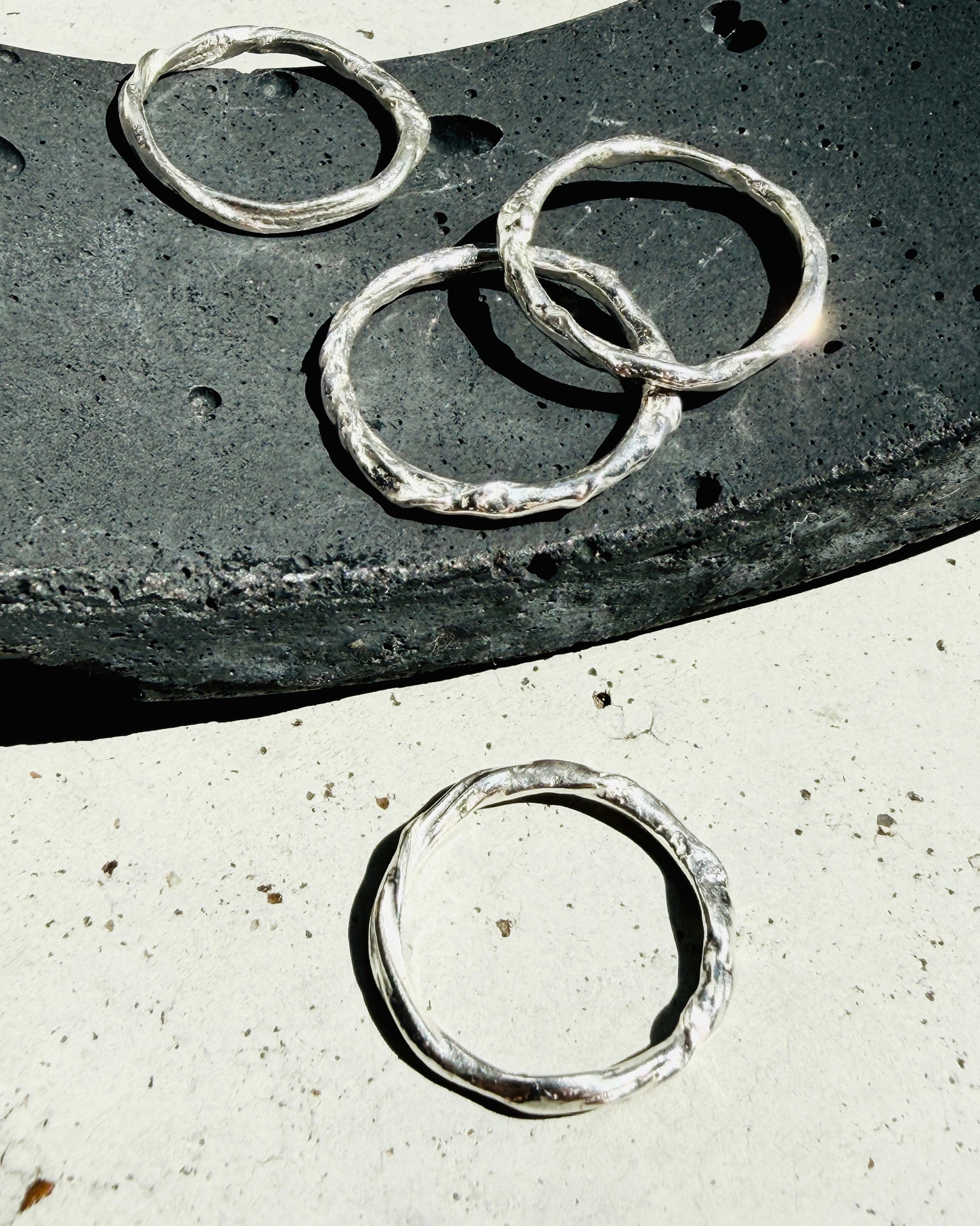 Three textured silver rings displayed on a dark stone slab and a light concrete surface under bright natural sunlight.
