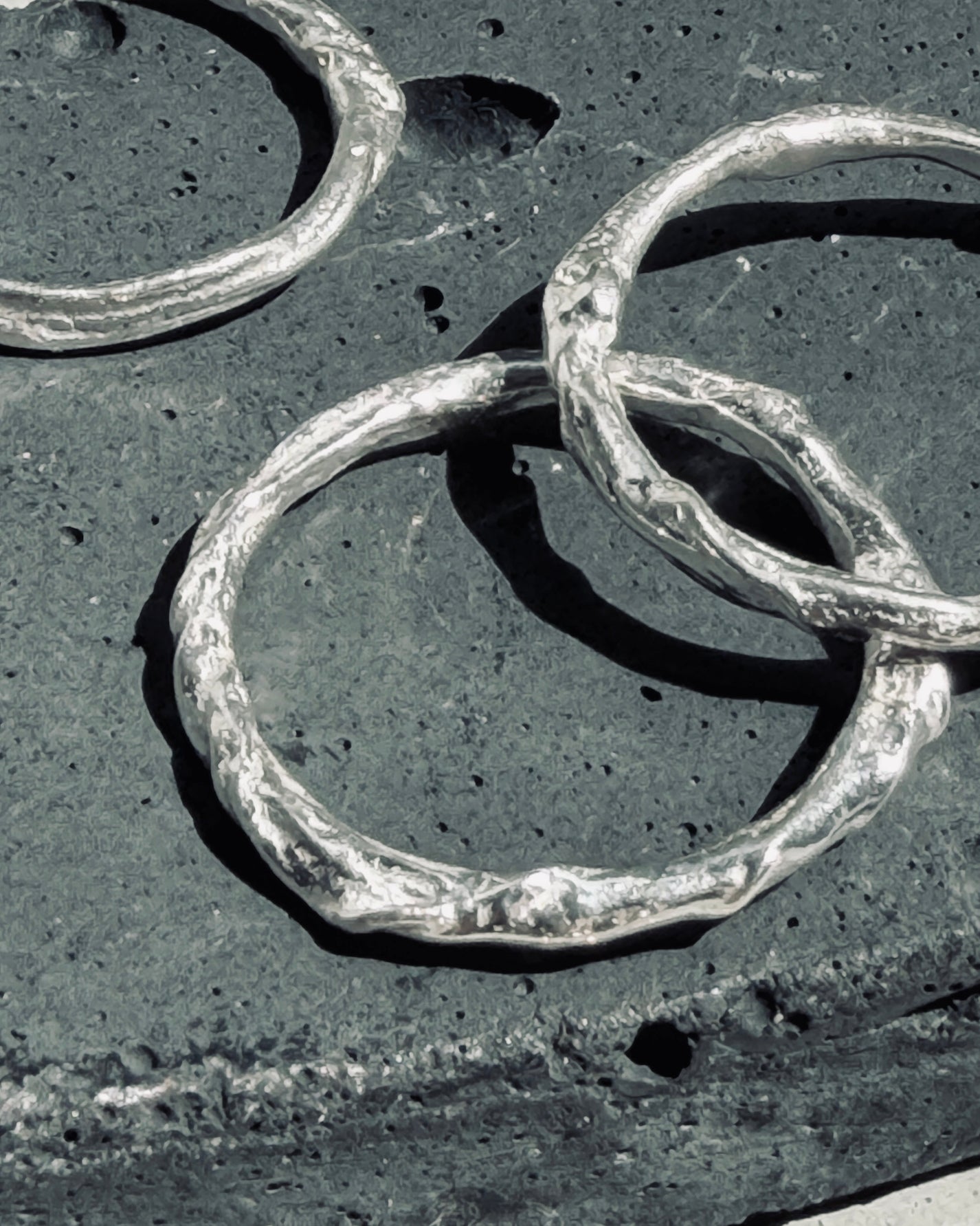 Three textured silver rings with organic, hammered edges overlapping on a dark charcoal stone surface.