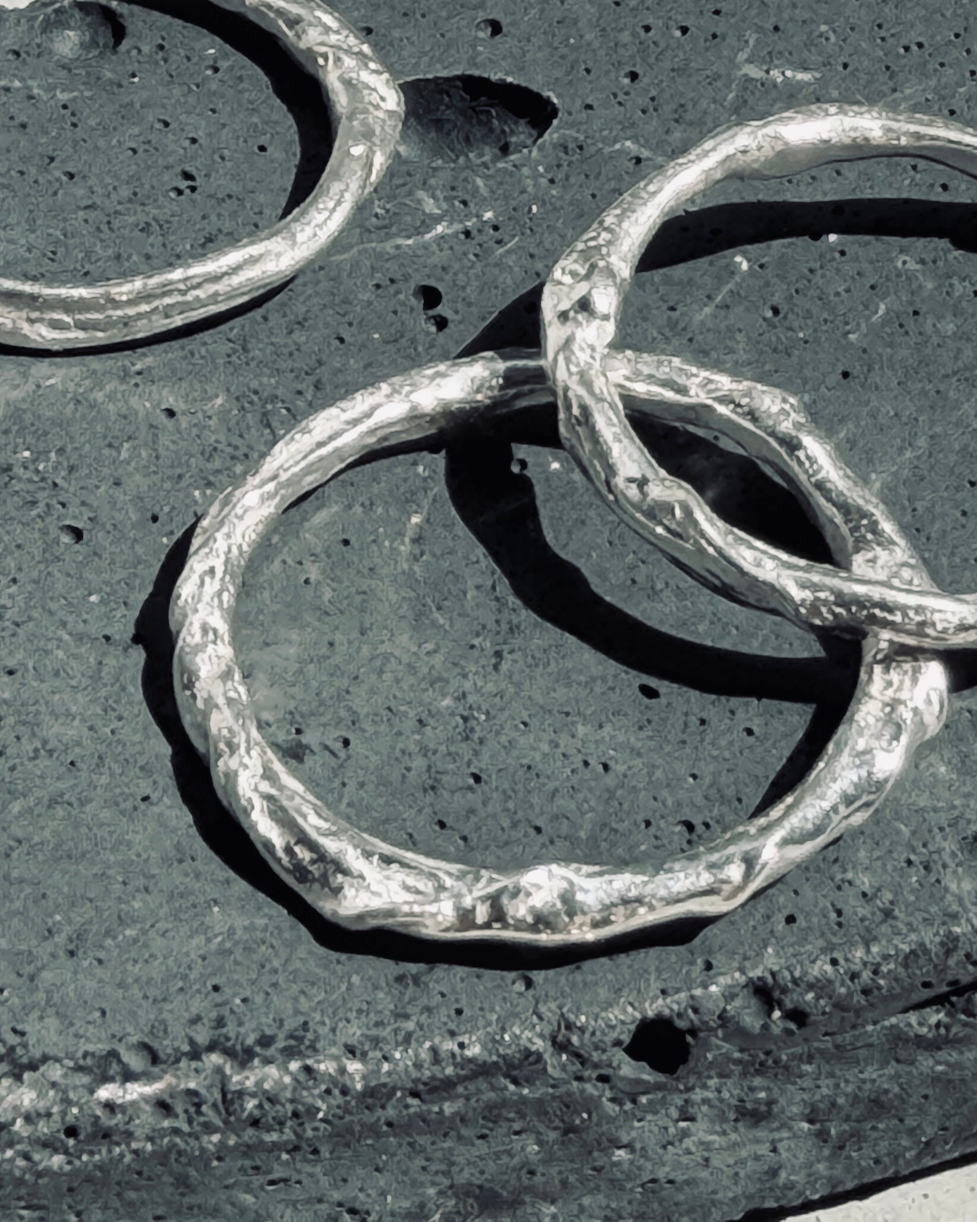 Three textured silver rings with organic, hammered edges overlapping on a dark charcoal stone surface.