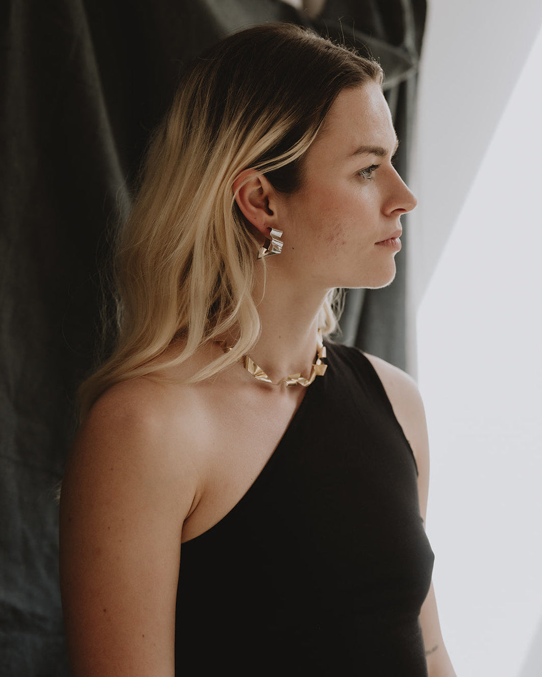 A woman in a black one-shoulder top wearing a gold chain necklace and matching gold drop earrings, shown in profile against a dark draped backdrop and bright window light.