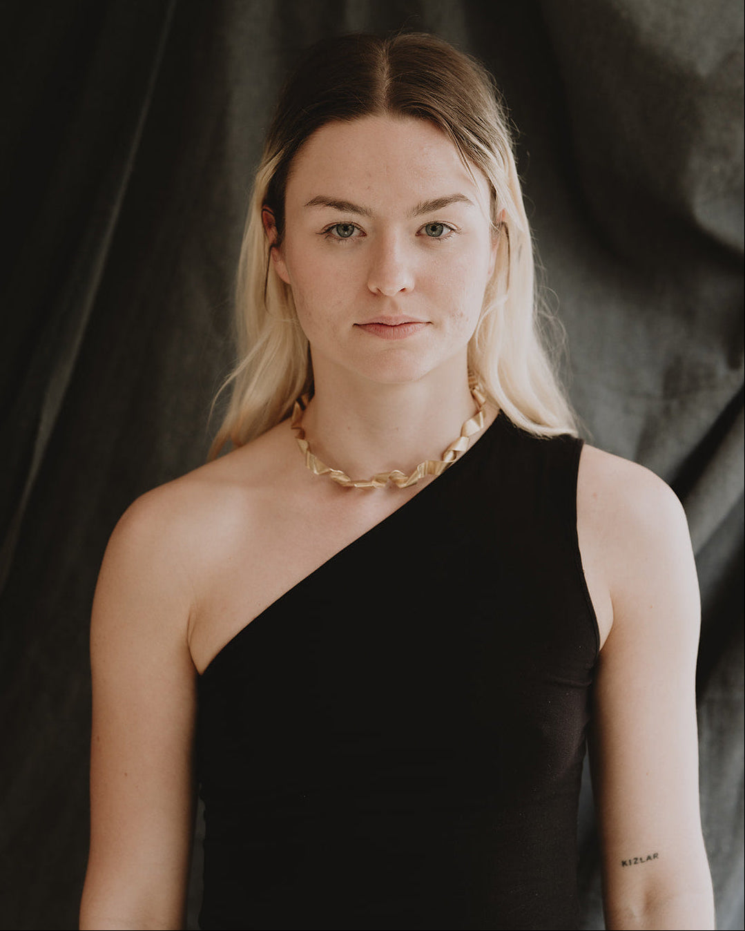 A blonde woman wearing a black one-shoulder top and a chunky gold chain necklace, standing against a dark draped fabric background.