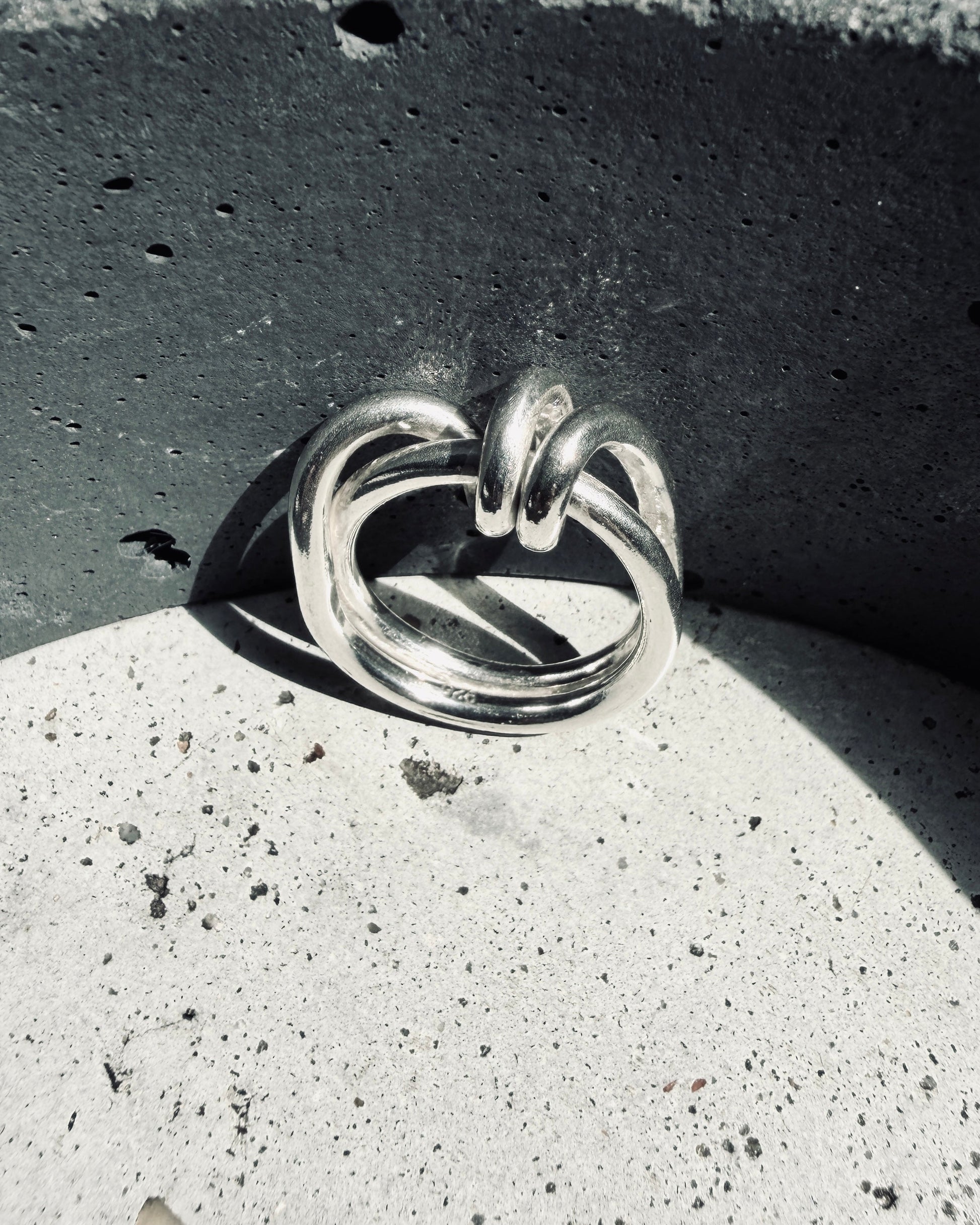 A polished silver ring with a knotted, interlocking band design, resting on a textured light gray concrete surface against a dark gray concrete background.