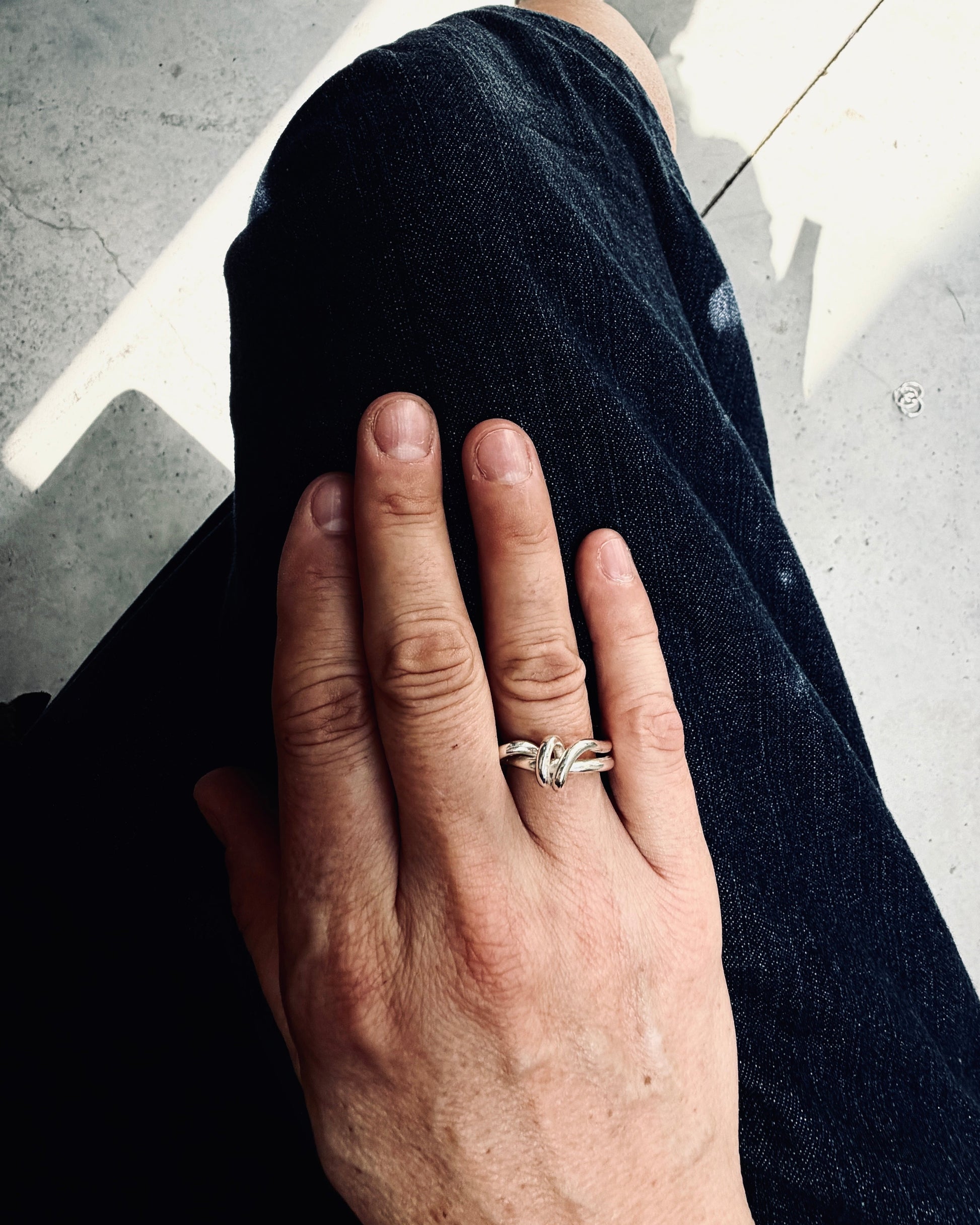 A close-up of a person's hand resting on their dark denim-clad leg, wearing a silver-toned double-band ring with a small clear gemstone.