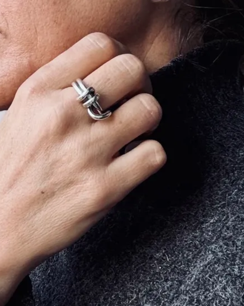 A close-up lifestyle shot of a person wearing a chunky silver ring on one finger and a textured silver hoop earring on the ear, with a dark gray knit sweater against a light gray background.