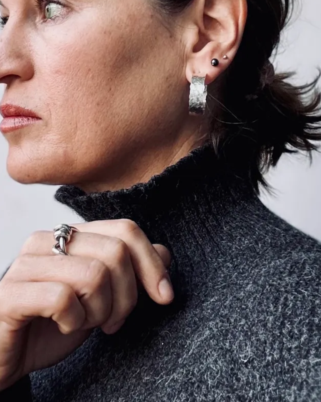 Close-up of a woman wearing a textured silver hoop earring and a silver ring, wearing a dark gray turtleneck sweater against a light gray background.