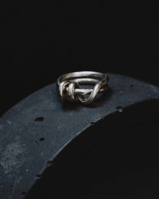 A silver ring with a knotted or intertwined band design, resting on the edge of a dark textured ceramic or stone arch against a black background.