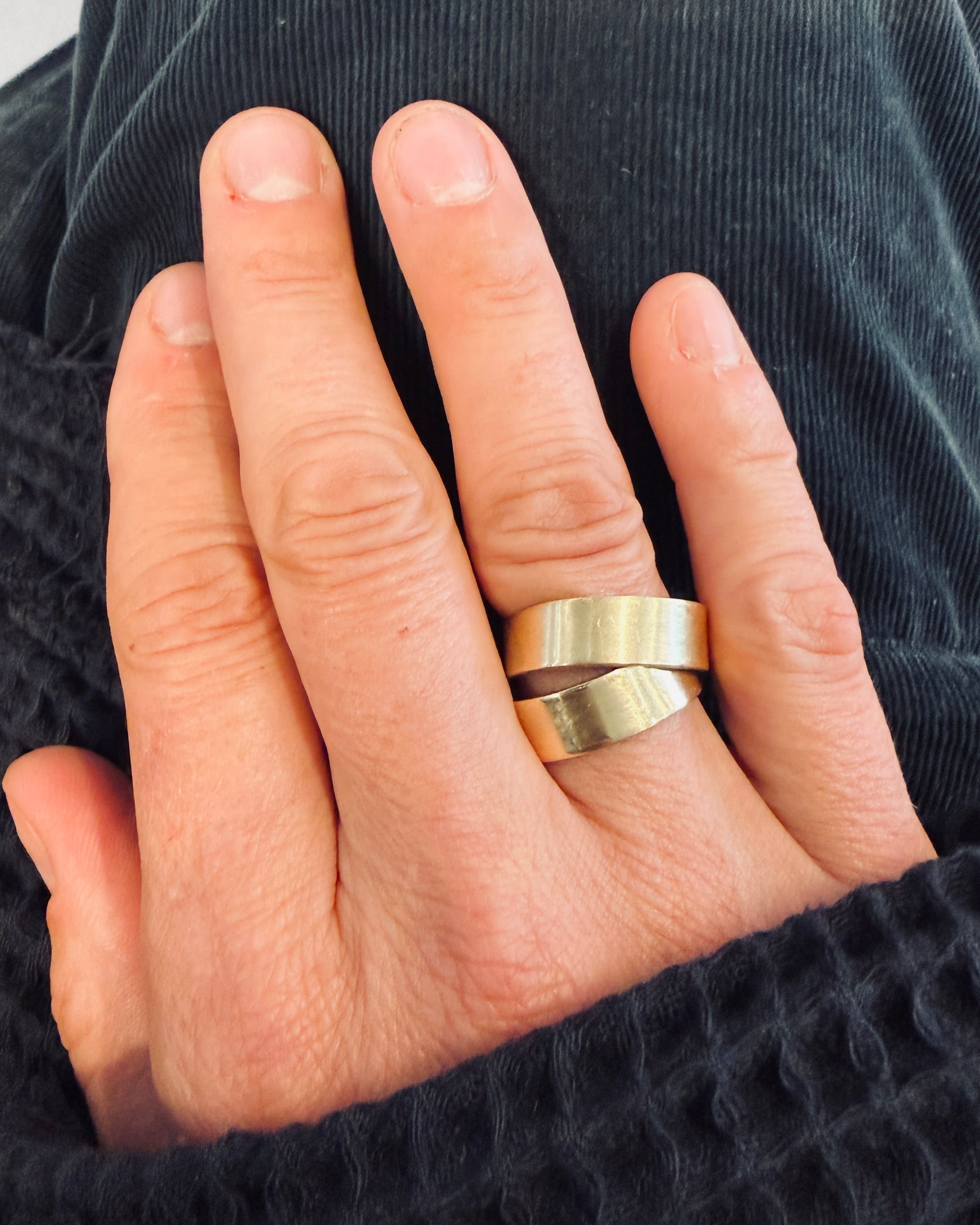 A close-up of a person's hand wearing two wide, polished gold bands, one flat and one with a wavy, brushed edge, set against a dark textured background.