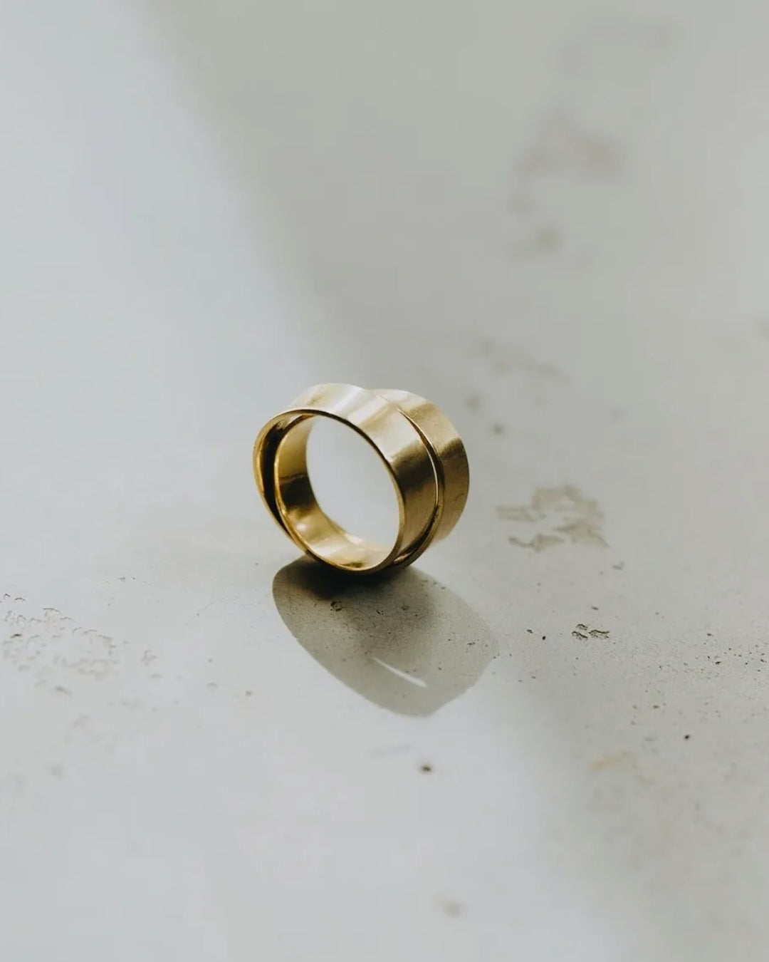 A gold-toned ring with a wide, chunky band and a central cutout, resting on a light gray surface with a soft reflection.