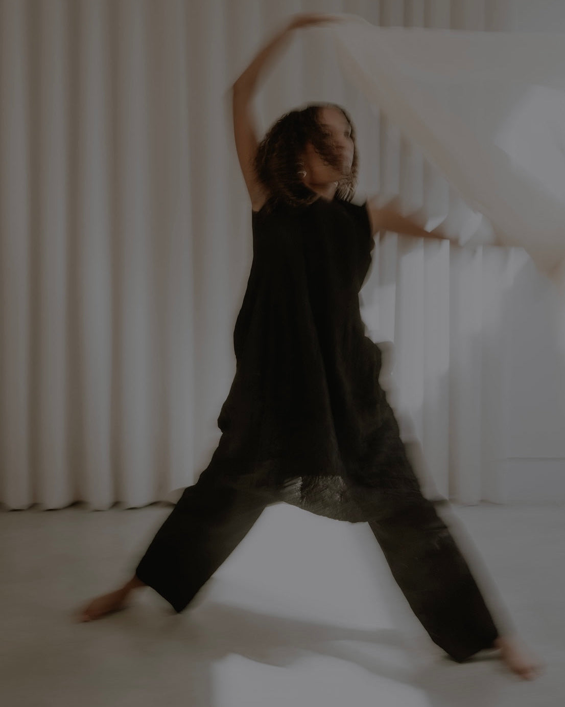 A woman in a loose black sleeveless top and wide-leg black pants poses dynamically in a bright studio with white curtains and sunlight streaming in.