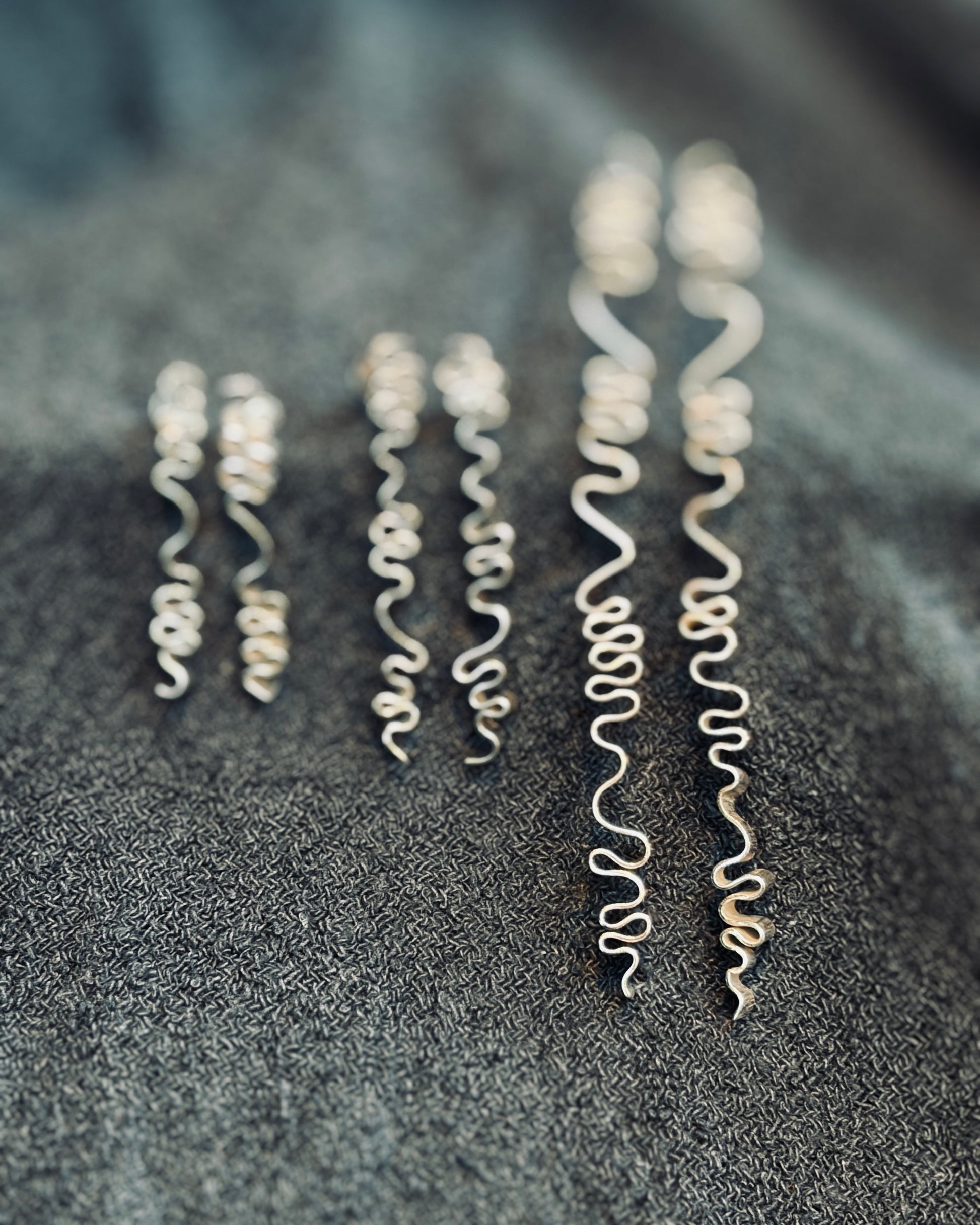 Five pairs of silver wavy spiral earrings arranged vertically on a dark textured fabric background.