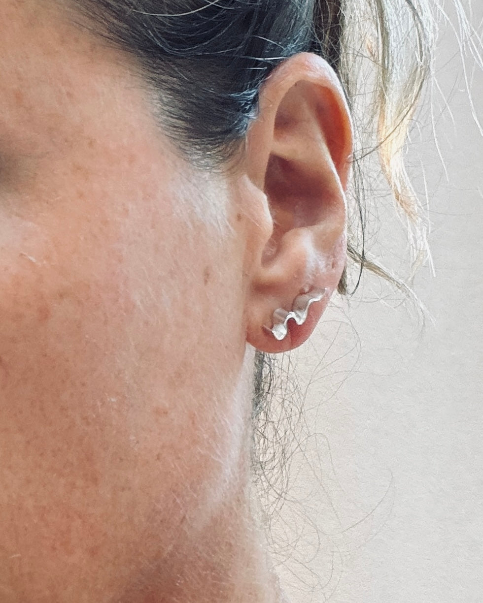 A close-up of a person's ear wearing a small silver-toned hoop earring with a wavy or scalloped front detail, against a plain light background.
