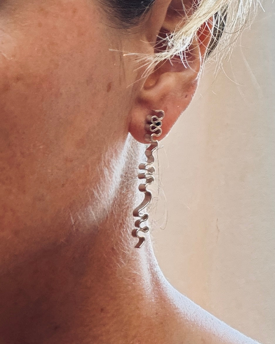 A close-up of a person's ear wearing a long, wavy, spiral-shaped dangling earring in a silver-tone metal.