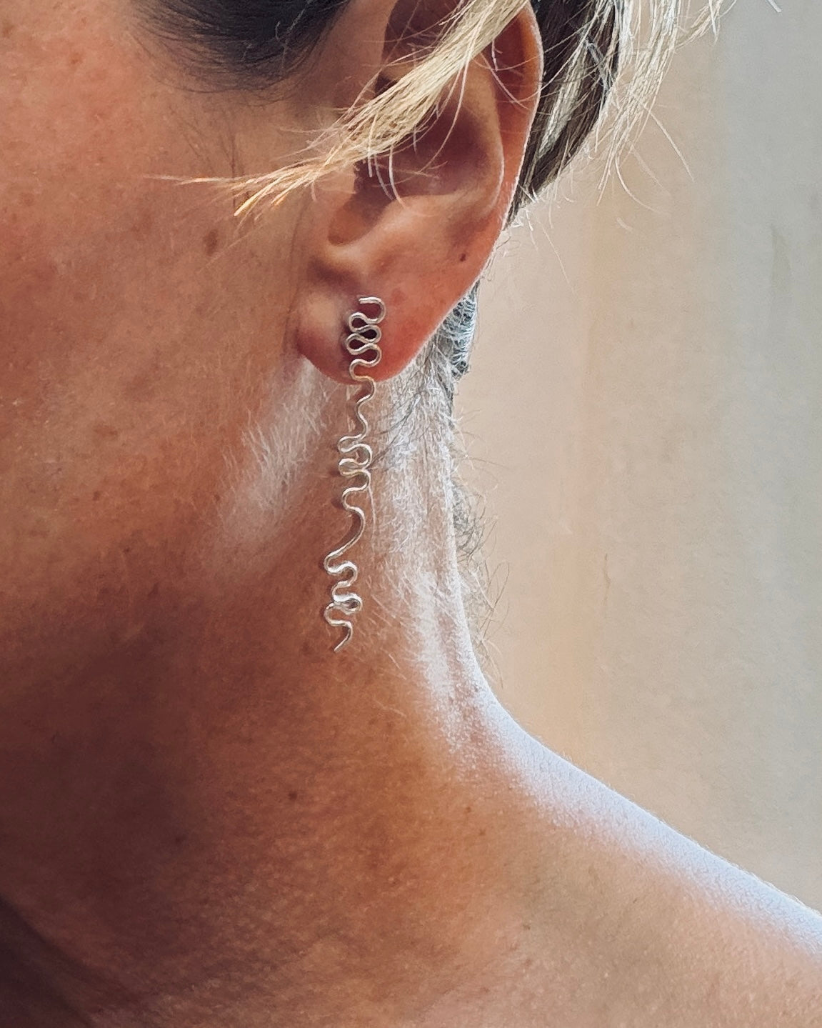A close-up of a person's ear wearing a long, delicate silver dangle earring with a wavy, coiled design.