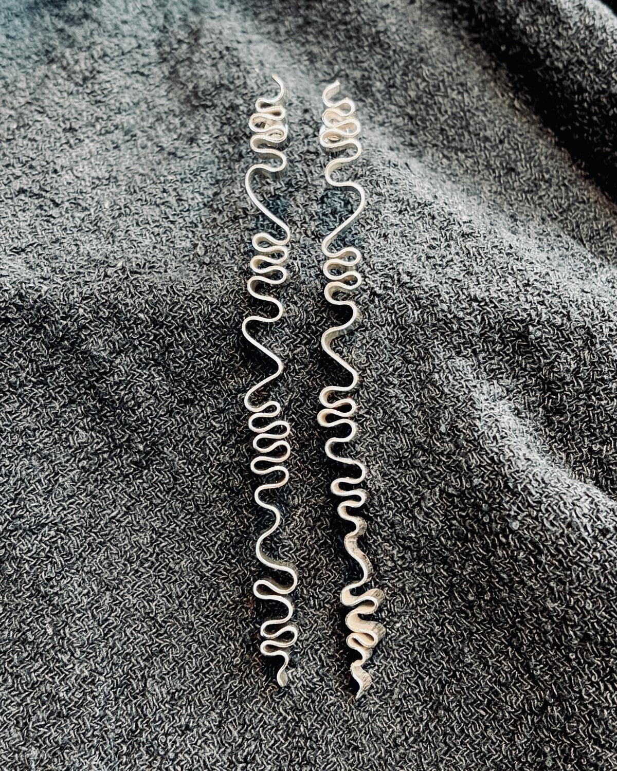 Two long, thin silver wavy strands laid flat on a dark gray textured fabric background.