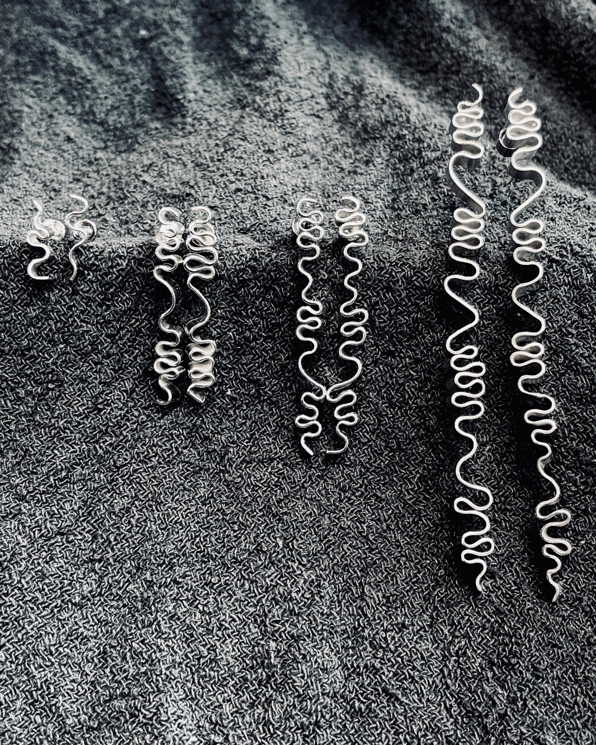 Five pairs of silver spiral drop earrings in varying sizes, arranged on a dark textured fabric background.