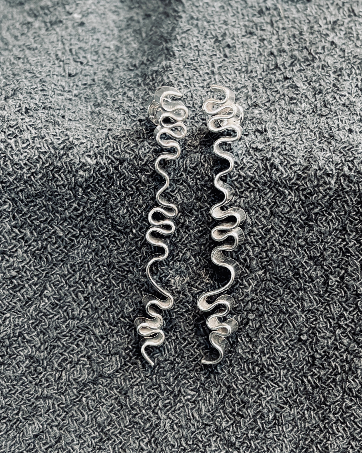A pair of long, wavy silver earrings laid flat on a textured dark gray fabric background.