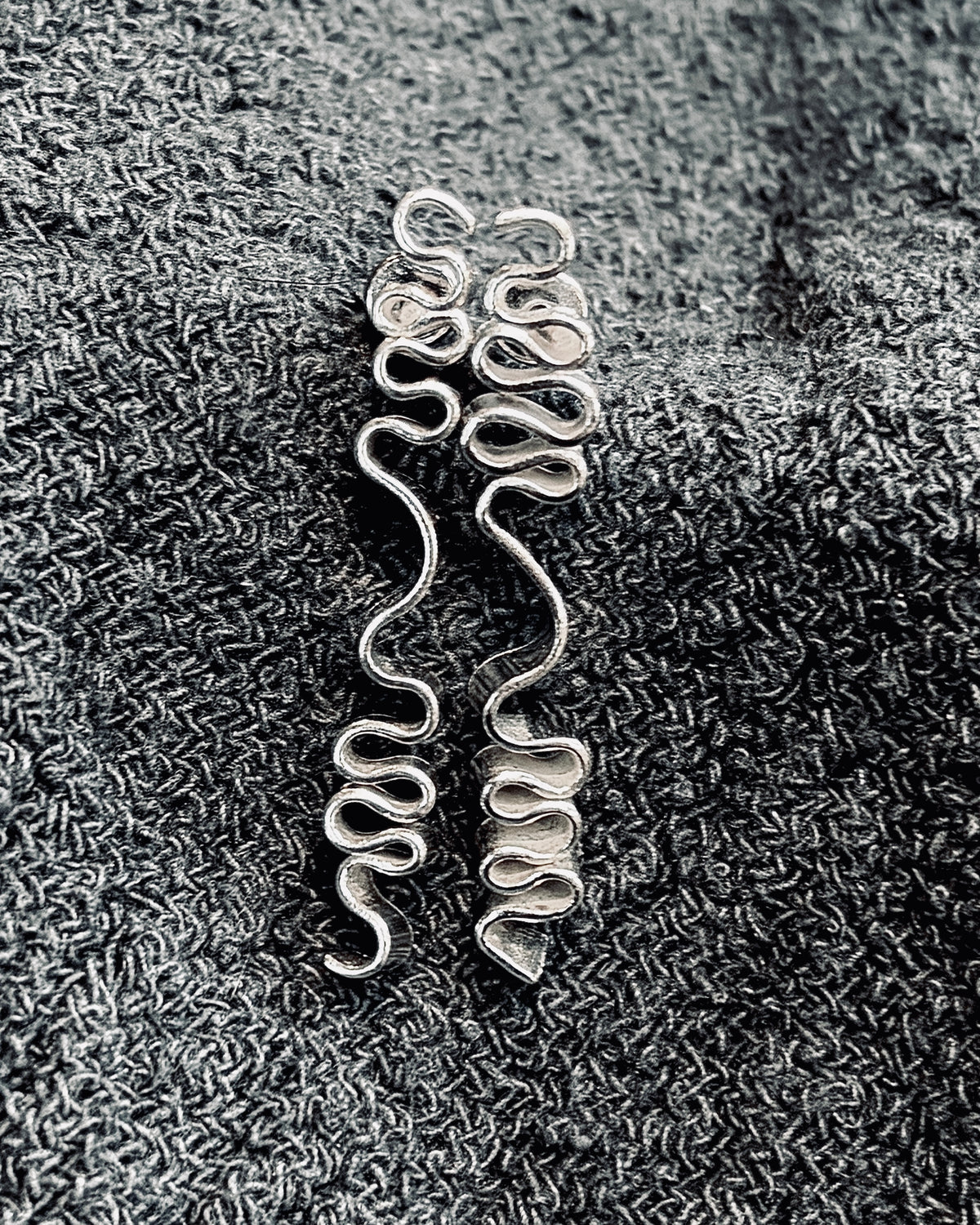 A pair of silver-toned spiral wire charms or earrings displayed on a textured dark gray fabric background.
