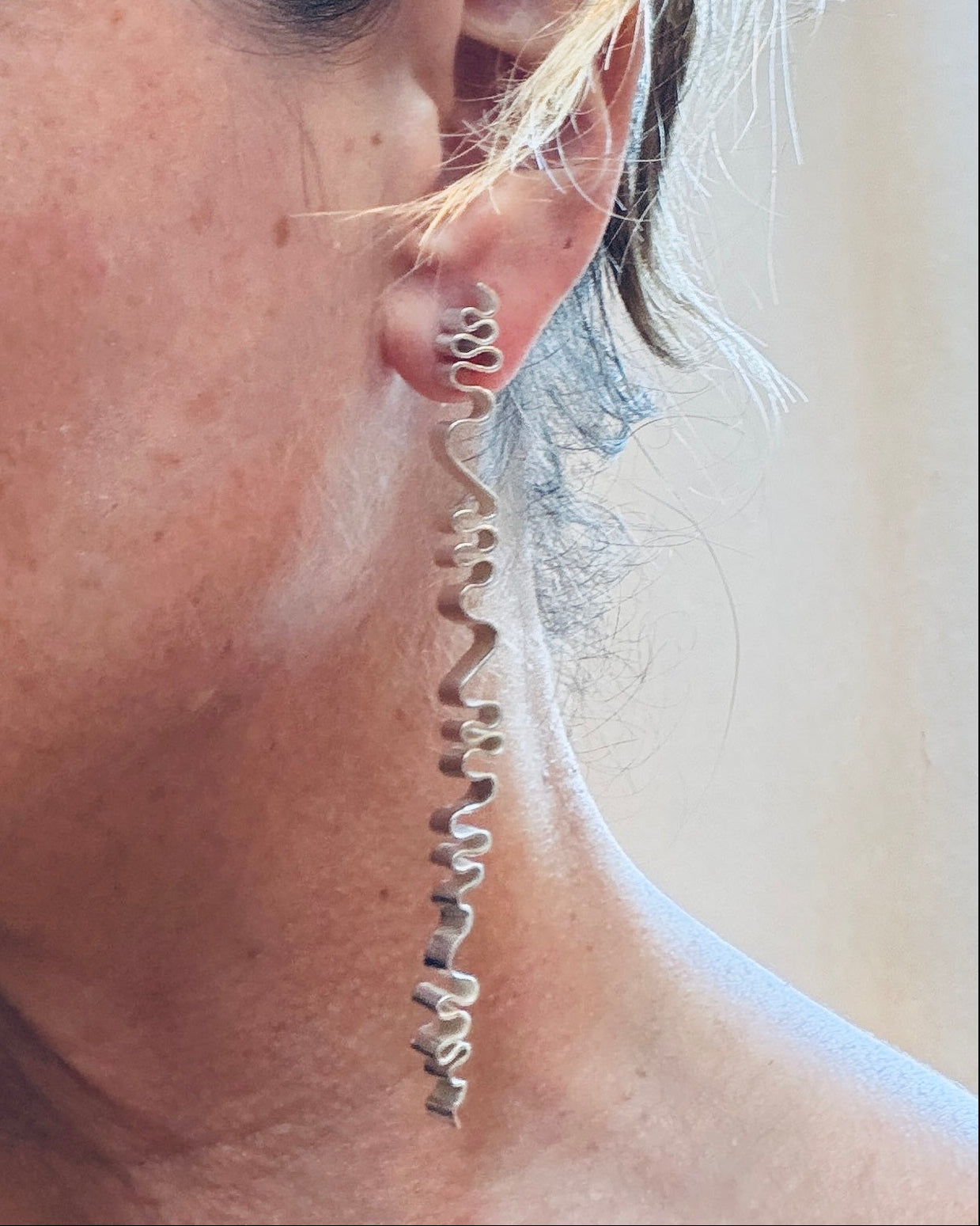 A close-up side profile of a woman wearing a long, wavy silver dangle earring that hangs from a small hoop earring.