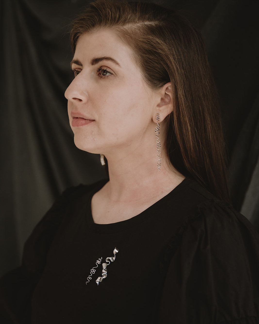 A woman with long brown hair is shown in profile against a dark draped backdrop, wearing a black top with a small white graphic on the chest and dangling earrings.