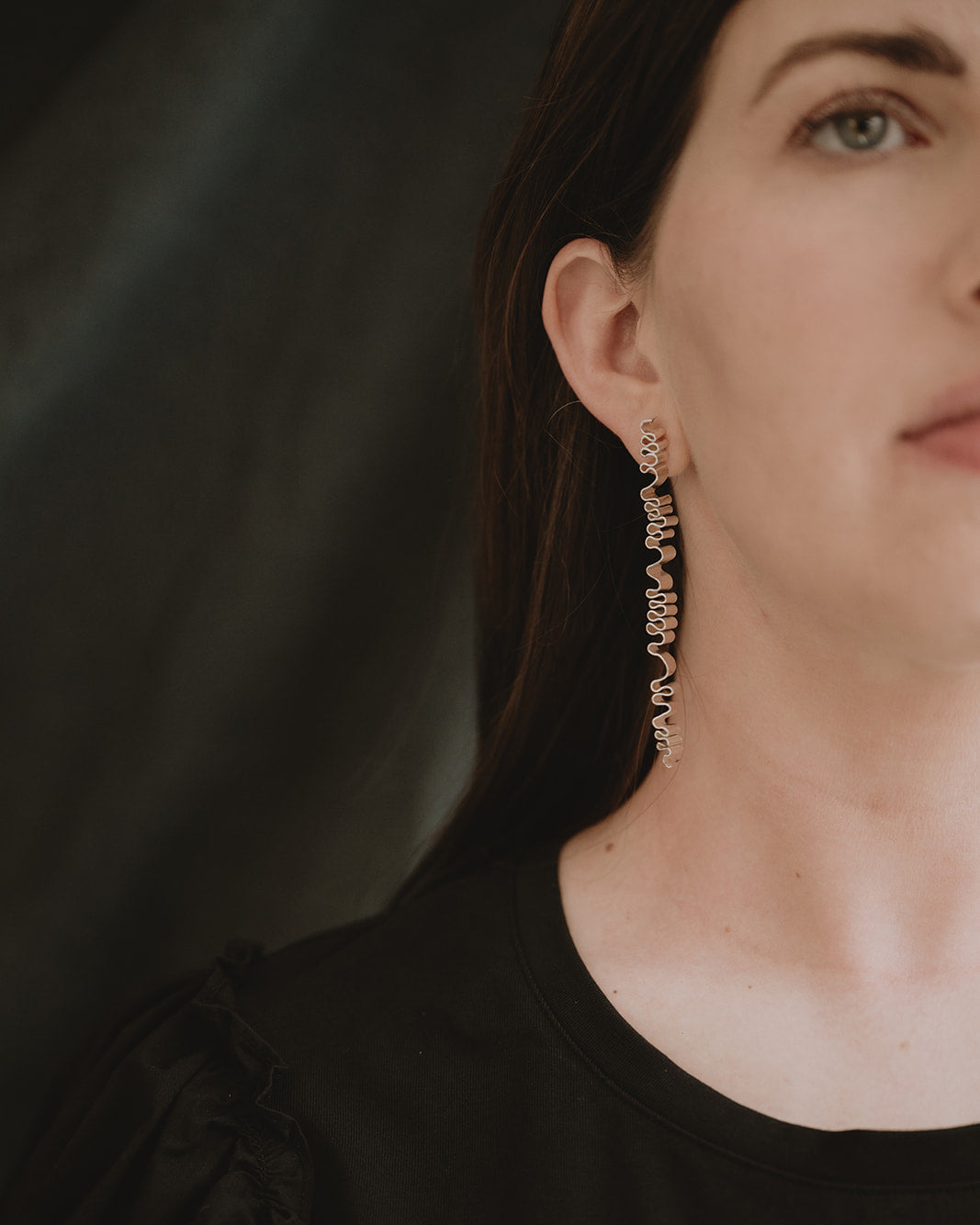 A close-up of a woman wearing a long, dangling wavy earring in a gold-tone metal setting, paired with a black top against a dark, draped background.