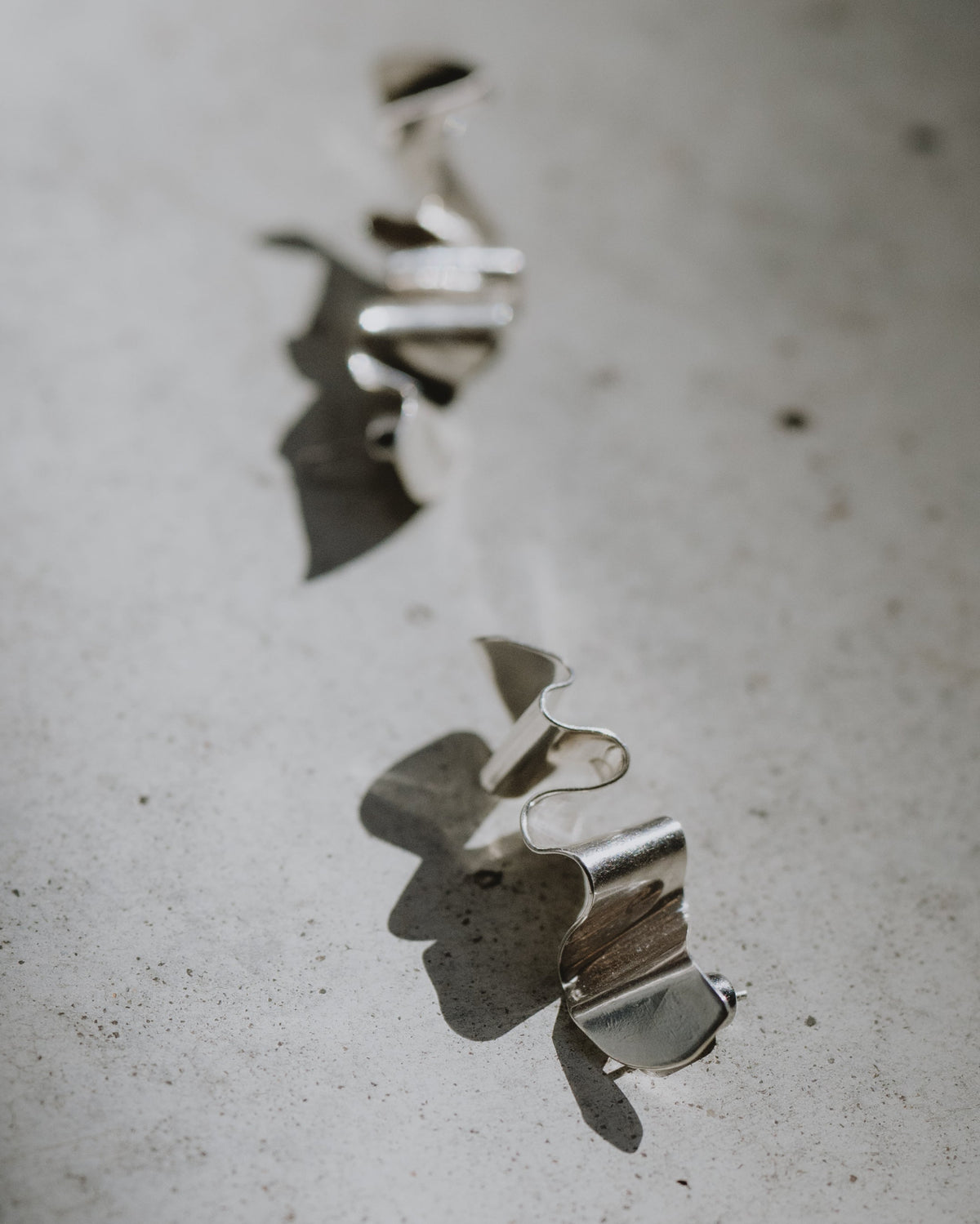 Two small silver metallic earring objects with curled edges resting on a textured light gray surface, with a diagonal shadow cast across the scene.