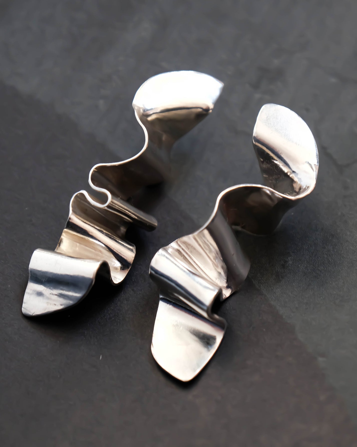 A pair of polished silver-toned metal earrings with organic, wavy, sculptural shapes, displayed on a dark textured surface.