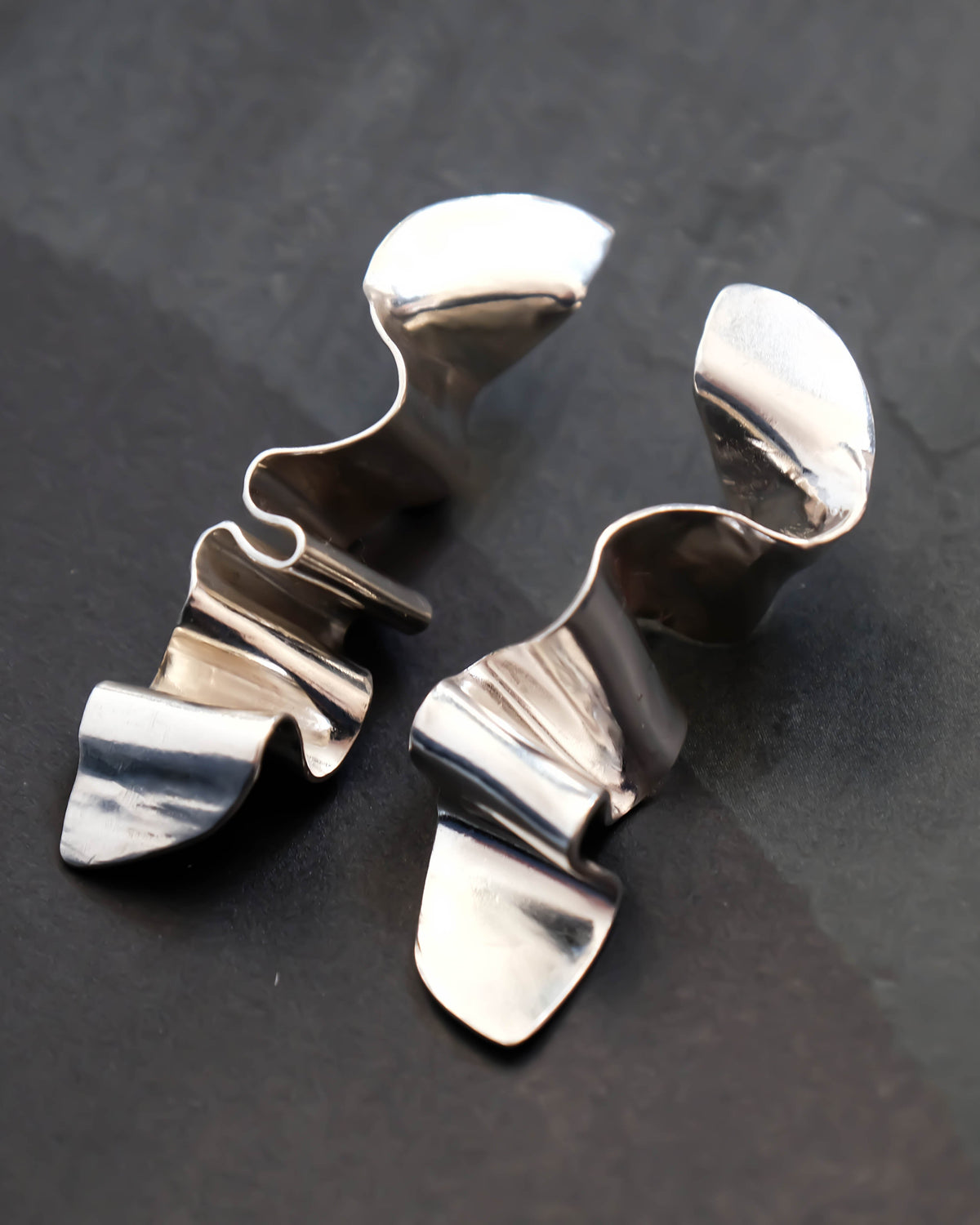 A pair of polished silver-toned metal earrings with organic, wavy, sculptural shapes, displayed on a dark textured surface.