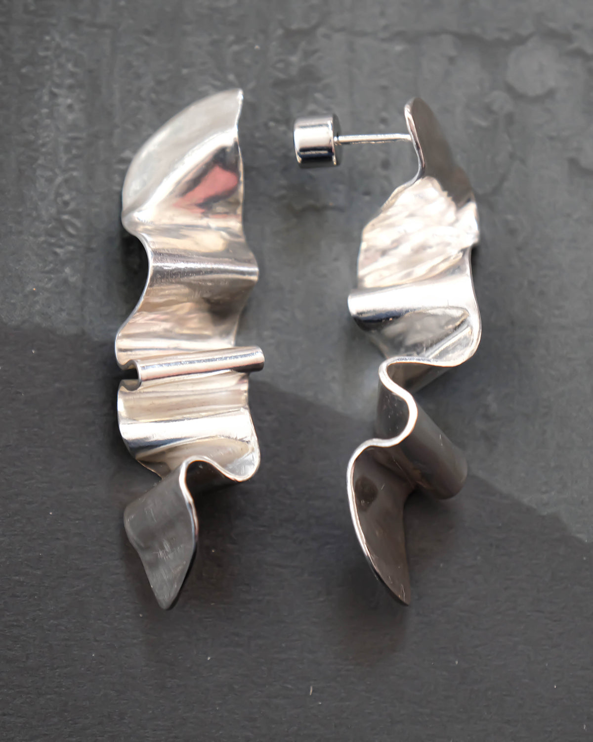 A pair of abstract, wavy sculptural earrings made of polished silver-tone metal, displayed on a dark textured slate surface.