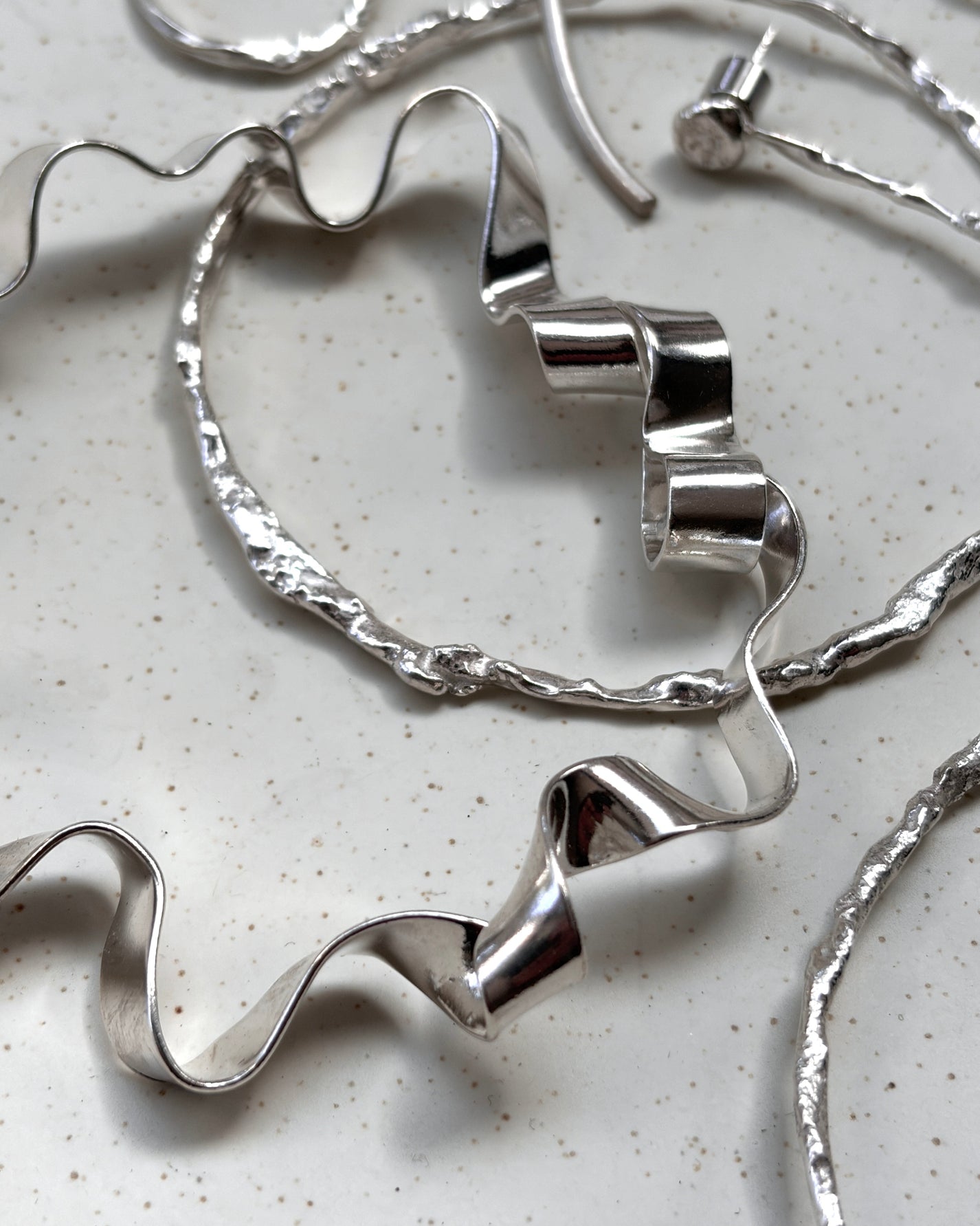 A close-up of silver-toned jewelry pieces arranged on a light speckled surface, including a wavy hoop and matching ribbon-like earrings or necklaces.