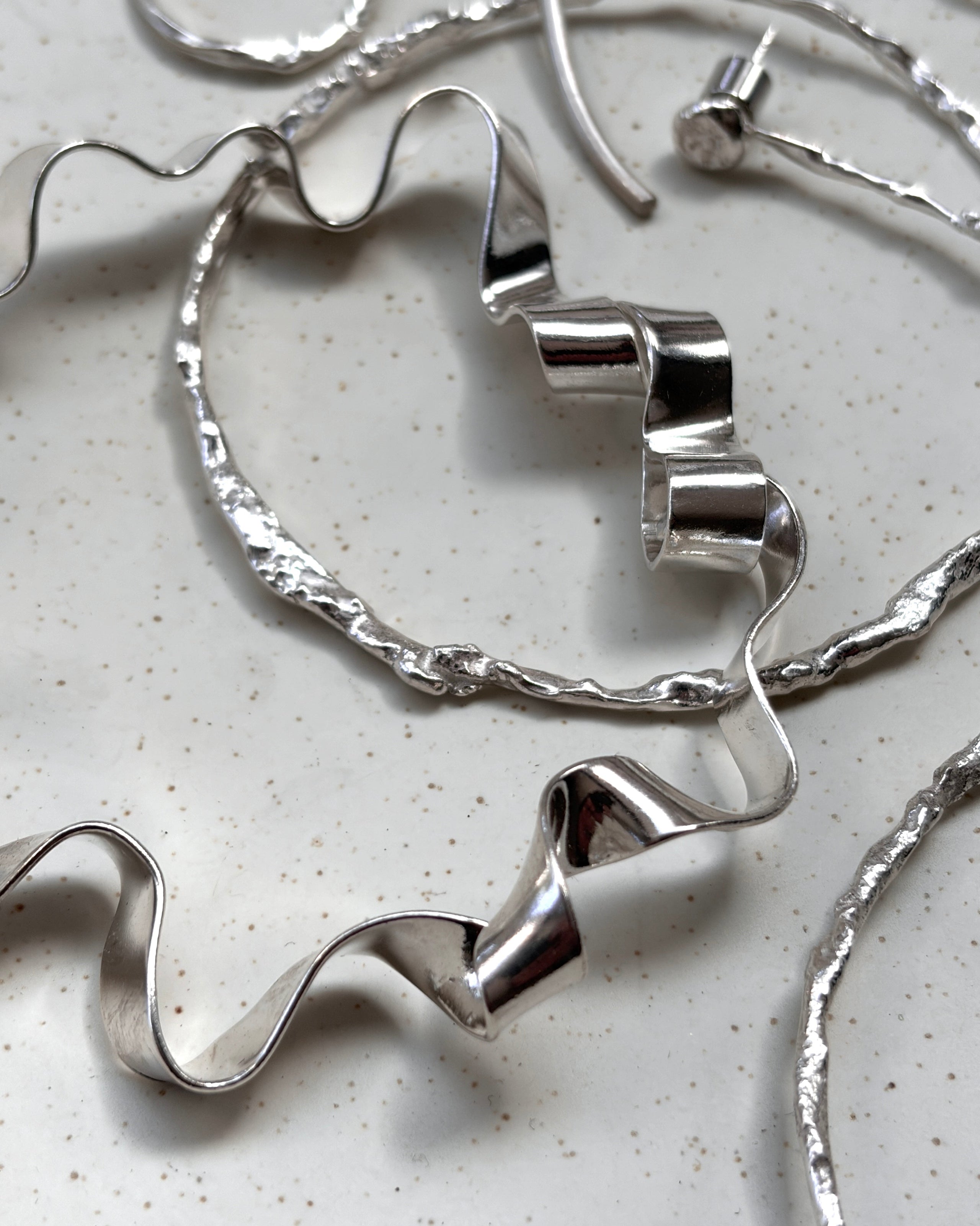 A close-up of silver-toned jewelry pieces arranged on a light speckled surface, including a wavy hoop and matching ribbon-like earrings or necklaces.