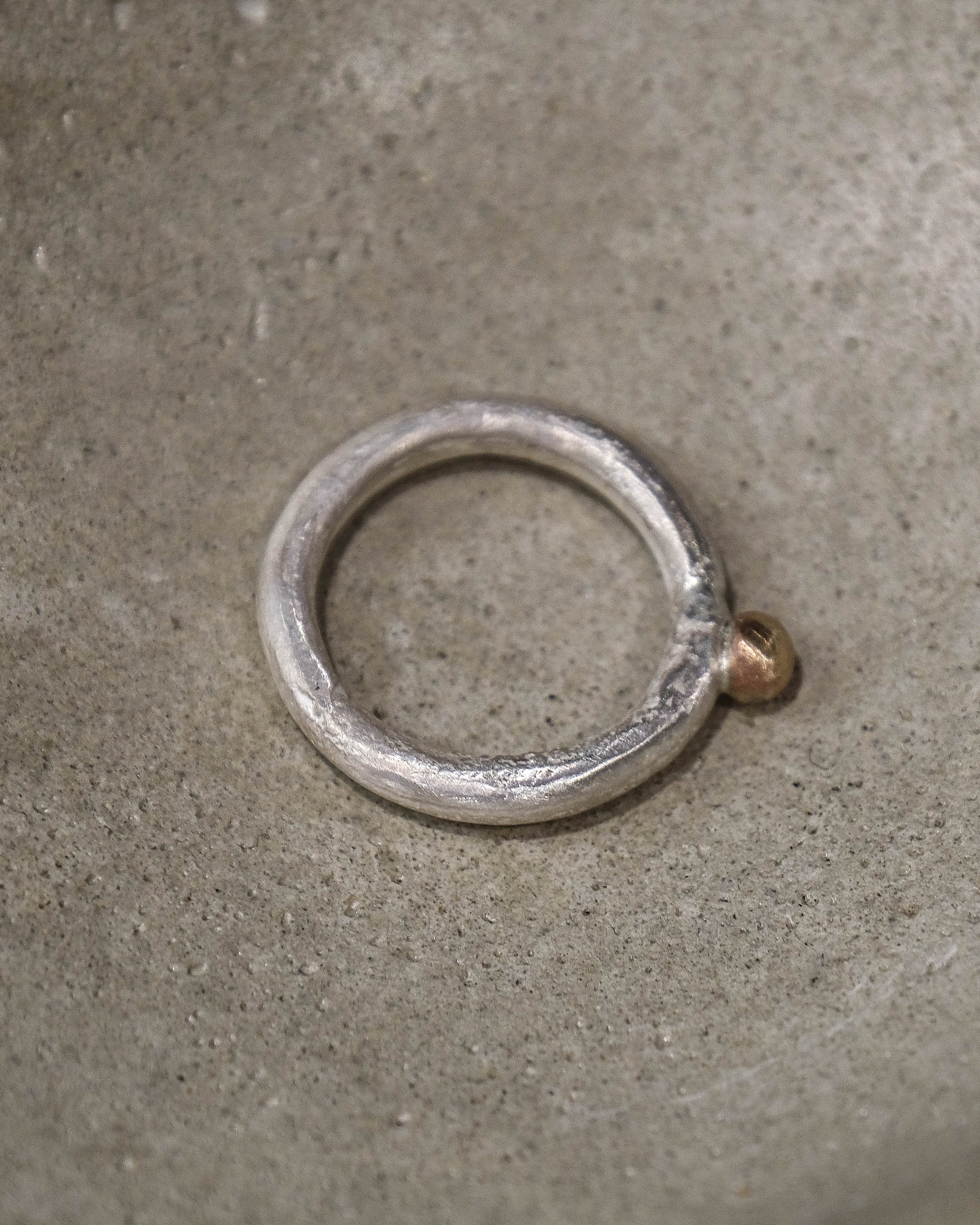 A silver-toned metal ring with a textured, hammered finish and a small brass-colored loop at the top, resting inside a textured gray concrete bowl.